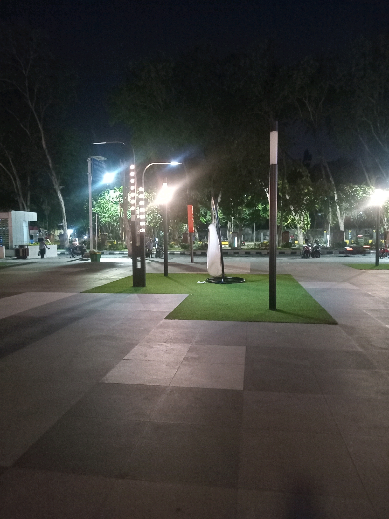 This image depicts a night scene in a public outdoor area, likely a park or pedestrian zone, in Sidoarjo, Indonesia. The foreground is dominated by a paved walkway with large, square stone tiles, casting long shadows from the artificial lighting. To the right and center, a section of artificial turf is laid out, with a tall, modern lamppost standing on its edge. In the midground, more lampposts illuminate a pathway that leads to a backdrop of trees. Several motorcycles are parked along the path. To the left, a person is walking away from the camera, dressed in dark clothing. Further back, a white, abstract sculpture stands on the turf. In the far left, a large, white structure with signage is visible, and a trash bin is located next to it. The overall ambiance is calm and illuminated by artificial light against the dark night sky.