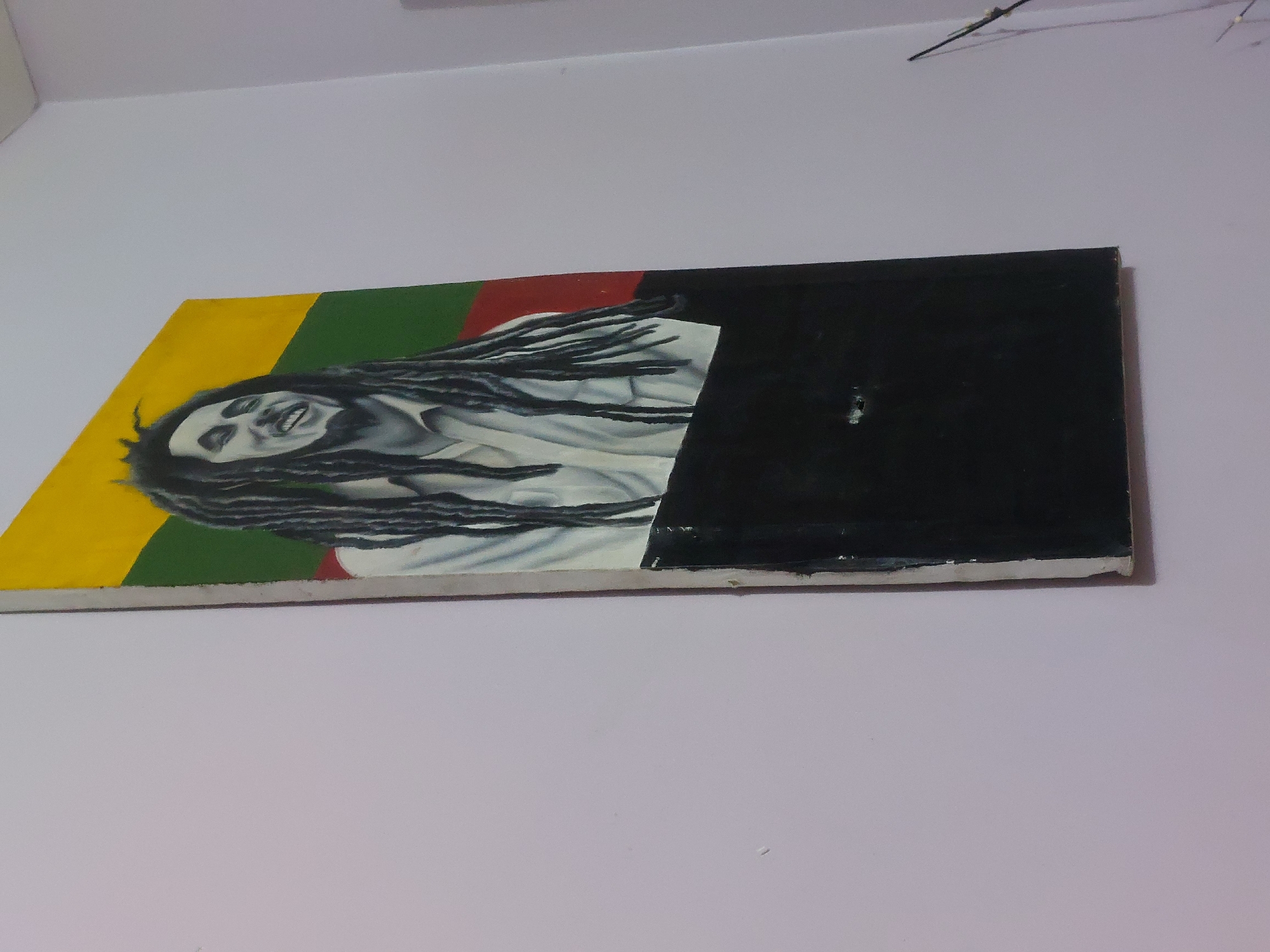 A painting of Bob Marley hangs on a wall. The painting is predominantly yellow, green, red, and black. Bob Marley's face and dreadlocks are painted on a lighter, neutral background. The location is unknown and is not discernible from the image.