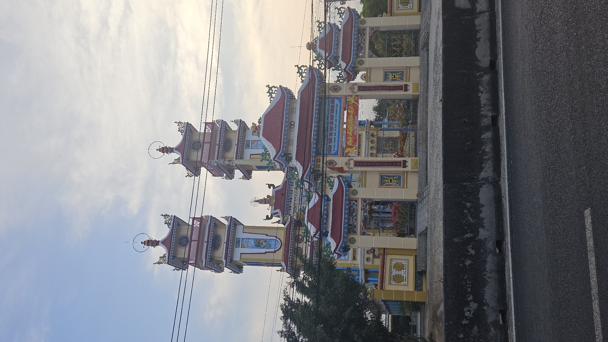This image depicts a religious site, potentially a Buddhist temple, in Cam Đức, Vietnam. The site features ornate architectural structures with red tiled roofs and yellow walls. The structures appear to be towers and gate structures, decorated with intricate details and religious iconography. Power lines are visible against the sky. The photograph is taken from a roadside. A concrete curb and asphalt road can be seen on the right side of the image. Trees are growing around the site.