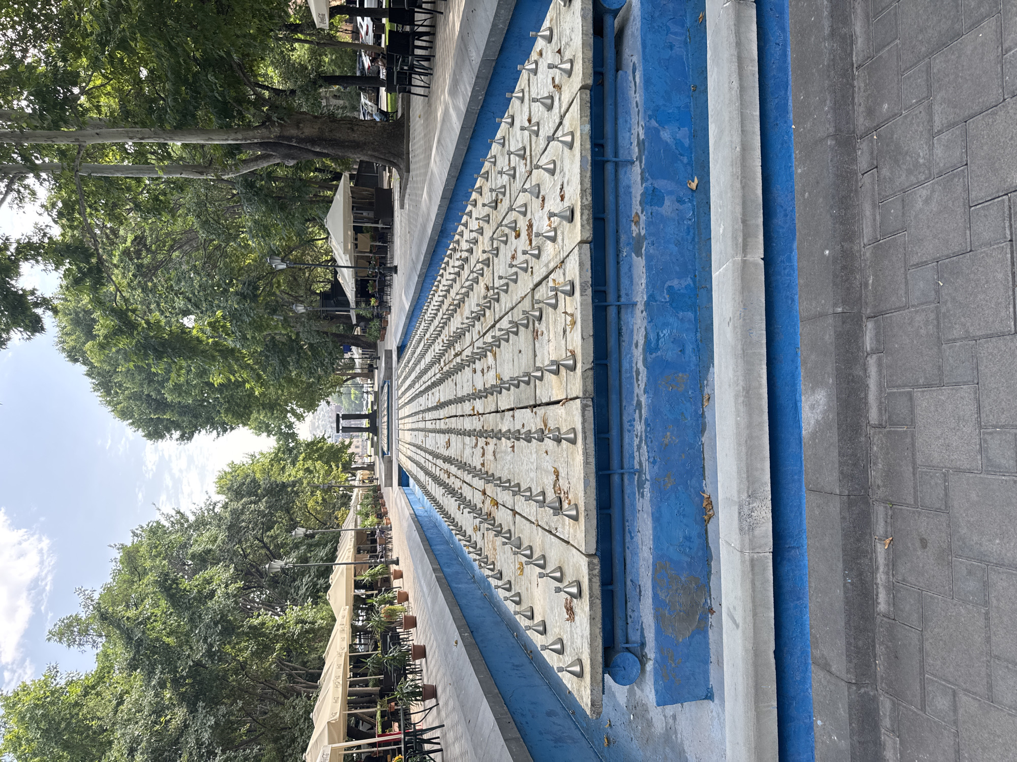 The image depicts a dry fountain in a park in Yerevan, Armenia. The fountain is a long, rectangular basin with a surface covered in rows of metal nozzles, likely for water jets. The basin itself is painted a vibrant blue, with some wear and tear visible. Surrounding the fountain are paved walkways and areas with outdoor seating, suggesting it is part of a public space or promenade. On either side of the fountain, trees provide shade, and in the background, there are more trees, buildings, and some parked cars. The sky is partly cloudy. No people are prominently featured in the immediate vicinity of the fountain. The overall scene suggests a warm, daytime setting in a city park.