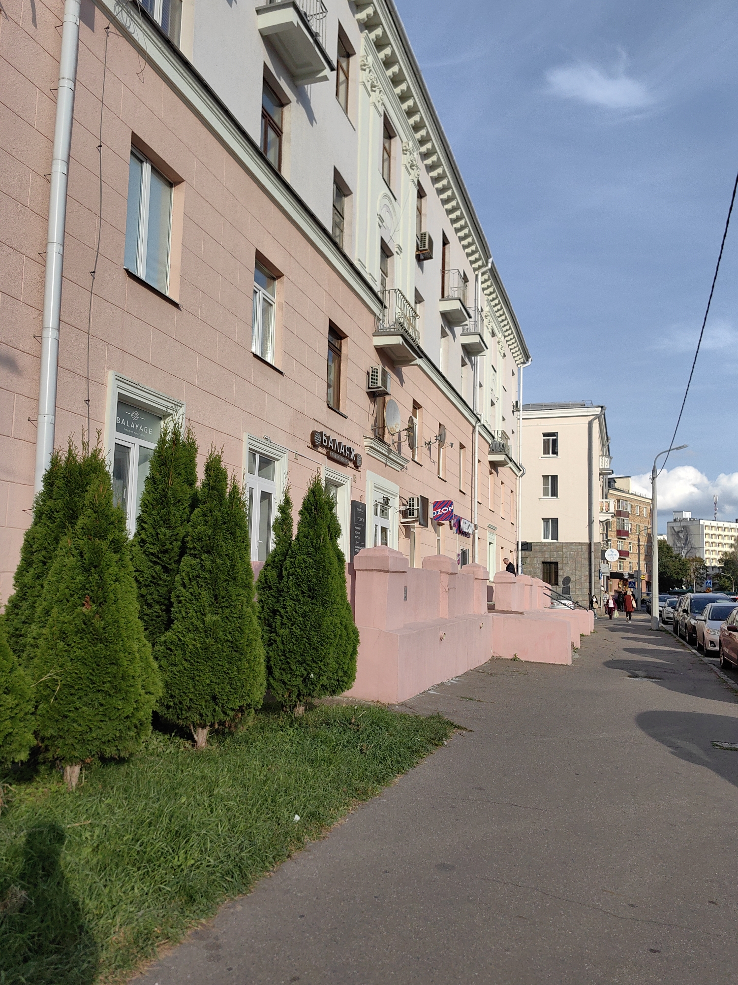The image shows a street in Minsk, Belarus, with a pink building on the left. The building has several windows and balconies, and there are trees and grass in front of it. A sidewalk runs along the street, and cars are parked on the right side. A few people are walking on the sidewalk. The sky is blue with some clouds. The building appears to be a commercial or residential building, with some shops on the ground floor. The overall impression is of a quiet urban scene on a sunny day.