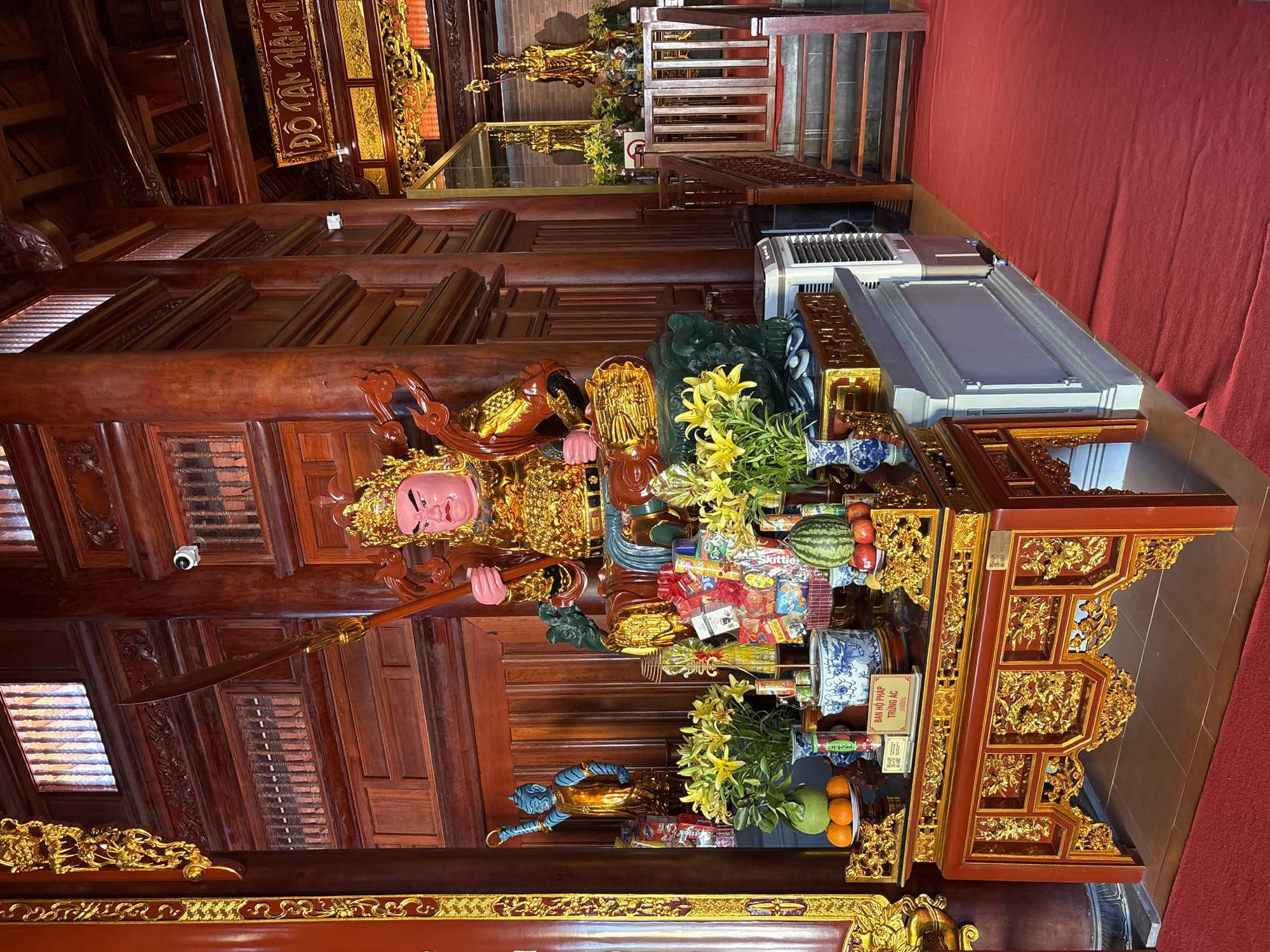 The image shows an interior scene in a temple, possibly located in Nam Nha Trang Ward, Vietnam. The temple features ornate wooden architecture, including carved panels and beams. A large, gilded statue of a figure with a red face and carrying a spear stands prominently. In front of the statue, there are offerings of flowers, fruit (including watermelons, oranges, and apples), vases, and packaged goods. The offerings are arranged on an intricately carved wooden table. The floor is covered with red carpeting. In the background, there are additional wooden structures and what appear to be stairs. An air conditioning unit is visible near the back. A security camera is also visible. The overall impression is one of richness, devotion, and reverence.