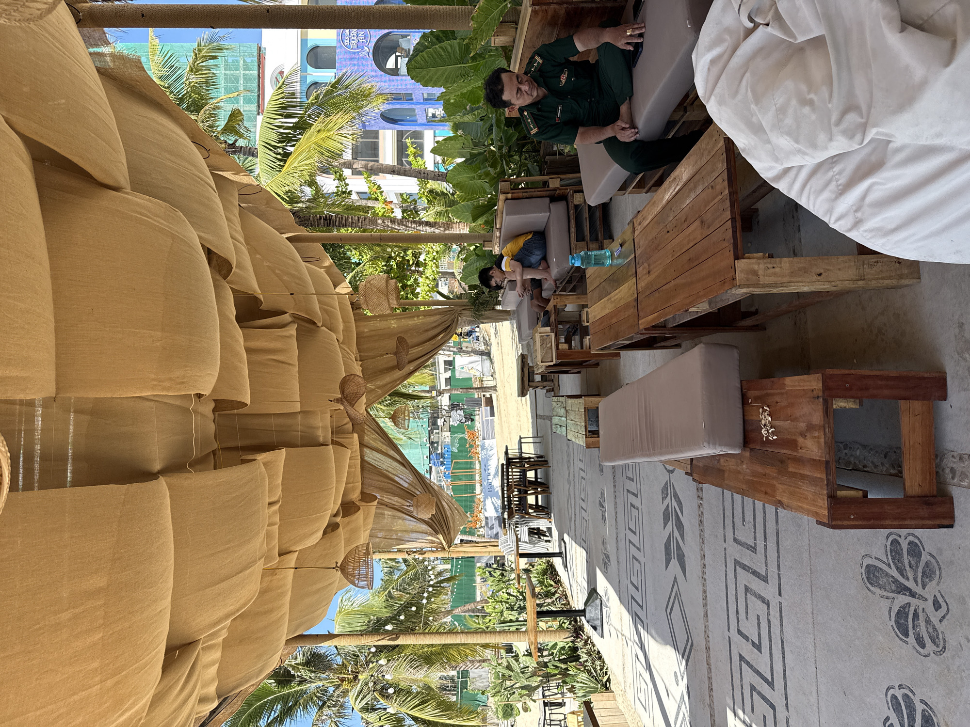 This media file captures an outdoor seating area, likely a cafe or restaurant, with a tropical vibe. The scene is set on a sunny day, with palm trees and lush greenery visible, suggesting a warm climate. The seating is rustic, with wooden furniture and comfortable cushions, some of which are piled up, creating a relaxed and informal atmosphere.
In the foreground, a large, textured tan fabric structure drapes over the seating area, providing shade. Further back, several tables and chairs are arranged, some occupied by people. Two individuals are clearly visible: a man in a green military-style uniform is seated on a couch, appearing to be looking at his phone. Nearby, a young child wearing a yellow shirt and blue shorts is crouched down, looking at something on the ground. In the background, a blue building with arched windows is visible, along with construction activity.
The ground is paved with a patterned surface, featuring geometric designs. Several light fixtures, some resembling woven lanterns, hang from the canopy. A bottle of water sits on one of the tables. The overall impression is one of a casual, open-air establishment in a sunny, possibly coastal, location. The presence of palm trees and the warm lighting indicate it's daytime. While there's no explicit text indicating the exact time, the bright sunlight suggests it's likely midday or early afternoon. The location context of Nha Trang, Vietnam, aligns with the visual cues of a tropical, beachside setting.
