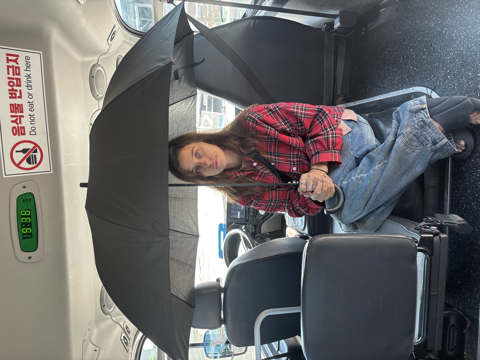This media file shows a young woman sitting in what appears to be the driver's seat of a vehicle, possibly a bus, given the interior view and the seats. She is holding a large, open black umbrella over herself, which obscures part of the interior and her face. The woman is wearing a red and black plaid shirt and blue jeans. Her left hand, adorned with rings, is holding the umbrella handle.
The scene is indoors, likely a vehicle's cabin. There are visible seats in the foreground and background. A digital display with green numbers is mounted on the left wall, showing "88:88". On the wall to the left of the display, there is a rectangular sign with red text and a circular prohibition symbol. The text on the sign is in Korean and English. The Korean text reads "음식물 반입금지" and the English text below it reads "Do not eat or drink here".
The lighting suggests it could be daytime, possibly overcast or rainy given the presence of the umbrella. The interior of the vehicle appears to be somewhat utilitarian. The overall mood is difficult to determine due to the angle and the umbrella, but the woman's direct gaze towards the camera might suggest a candid moment or an awareness of being photographed. The context provided indicates the location is Jeju, South Korea.
