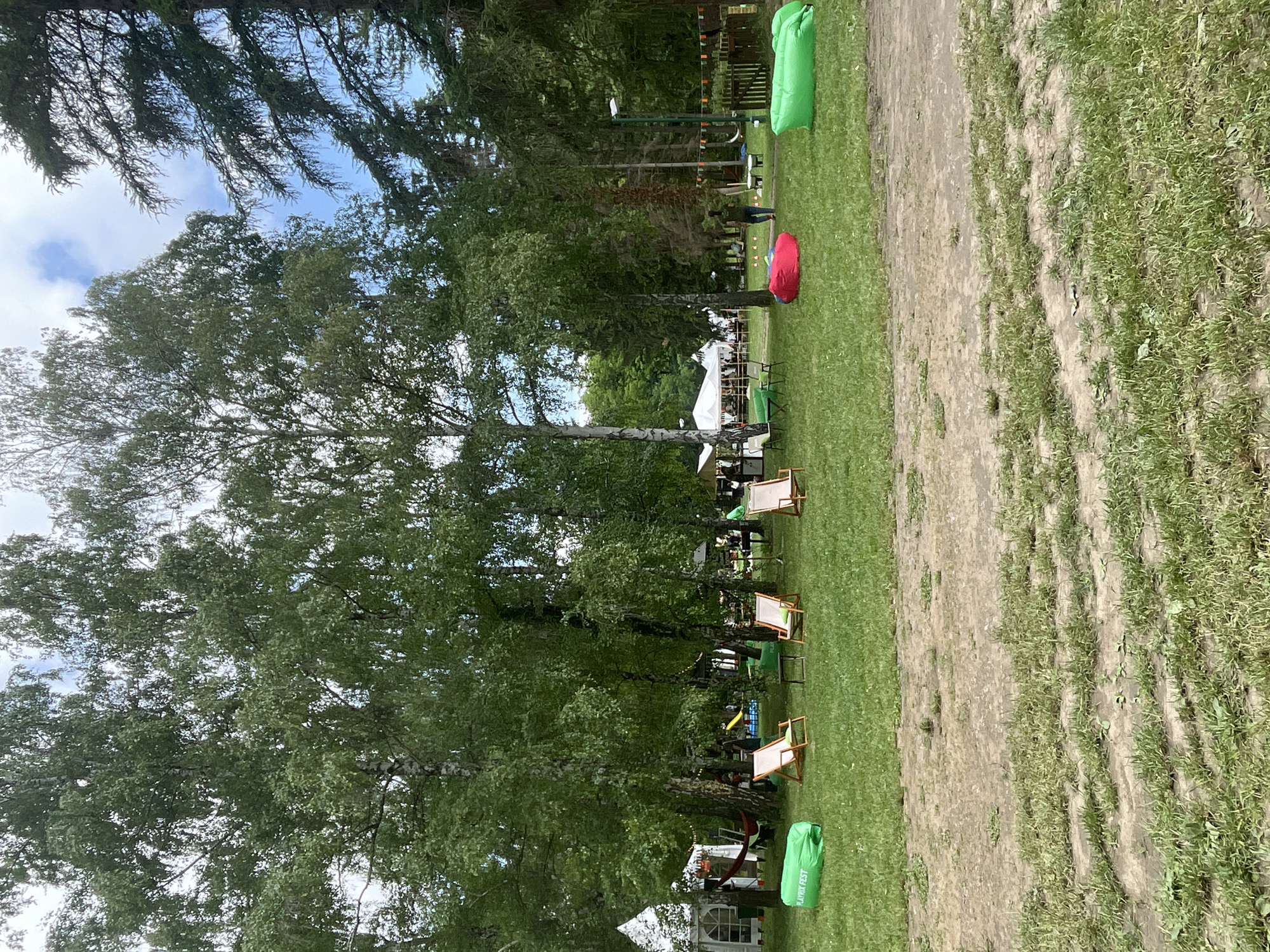 This image captures an outdoor recreational area within Sremski Karlovci Municipality, Serbia, on a partly cloudy day. The scene is dominated by numerous trees, primarily birches with light-colored bark and dense green foliage, interspersed with darker conifers on the right side, framing a bright blue sky with white clouds. The ground consists of a grassy lawn showing significant wear with patches of exposed dirt and discernible tracks in the foreground and midground.
In the immediate foreground, a bright green inflatable lounge bag marked "PLAYRIX FEST" is positioned on the left. To its right, three wooden-framed deck chairs with light-colored canvas seats are arranged around a small, dark, round table. Further right, a red inflatable lounge bag and another green inflatable lounge bag are visible on the grass.
In the midground and background, several white temporary tent structures are set up, indicating an event or gathering. On the far left, a white peaked tent with clear window panels is visible, near which a red hammock is strung between two trees. Further back, larger white marquee tents are present, with indistinct figures of people moving around them, suggesting activity. A yellow slide is faintly visible in the deeper background, near the center. On the right side, a person wearing a dark top and lighter trousers is walking away from the viewer, towards the right. A string of colorful triangular pennant banners (orange, green, red) is strung between trees and a light pole on the far right, adding a festive element to the scene. The overall impression is that of a temporary outdoor event in a natural park-like setting.