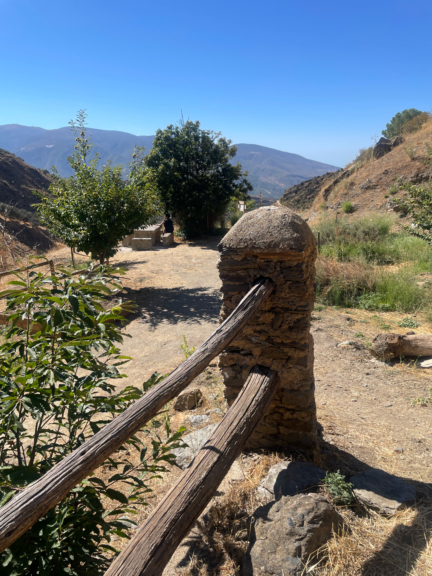 The image depicts an outdoor scene in Lanjarón, Spain, under a clear blue sky. In the foreground, a rustic stone pillar supports a weathered wooden fence railing. To the left, lush green foliage of a young tree contrasts with the dry, sun-baked ground. In the middle ground, a person is seated on a stone bench next to a stone picnic table, shaded by a mature tree. Beyond this area, rolling hills and mountains recede into the distance. The terrain is characterized by dry grass, scattered rocks, and sparse vegetation, indicative of a warm, arid climate. The overall impression is one of a tranquil, rural setting.