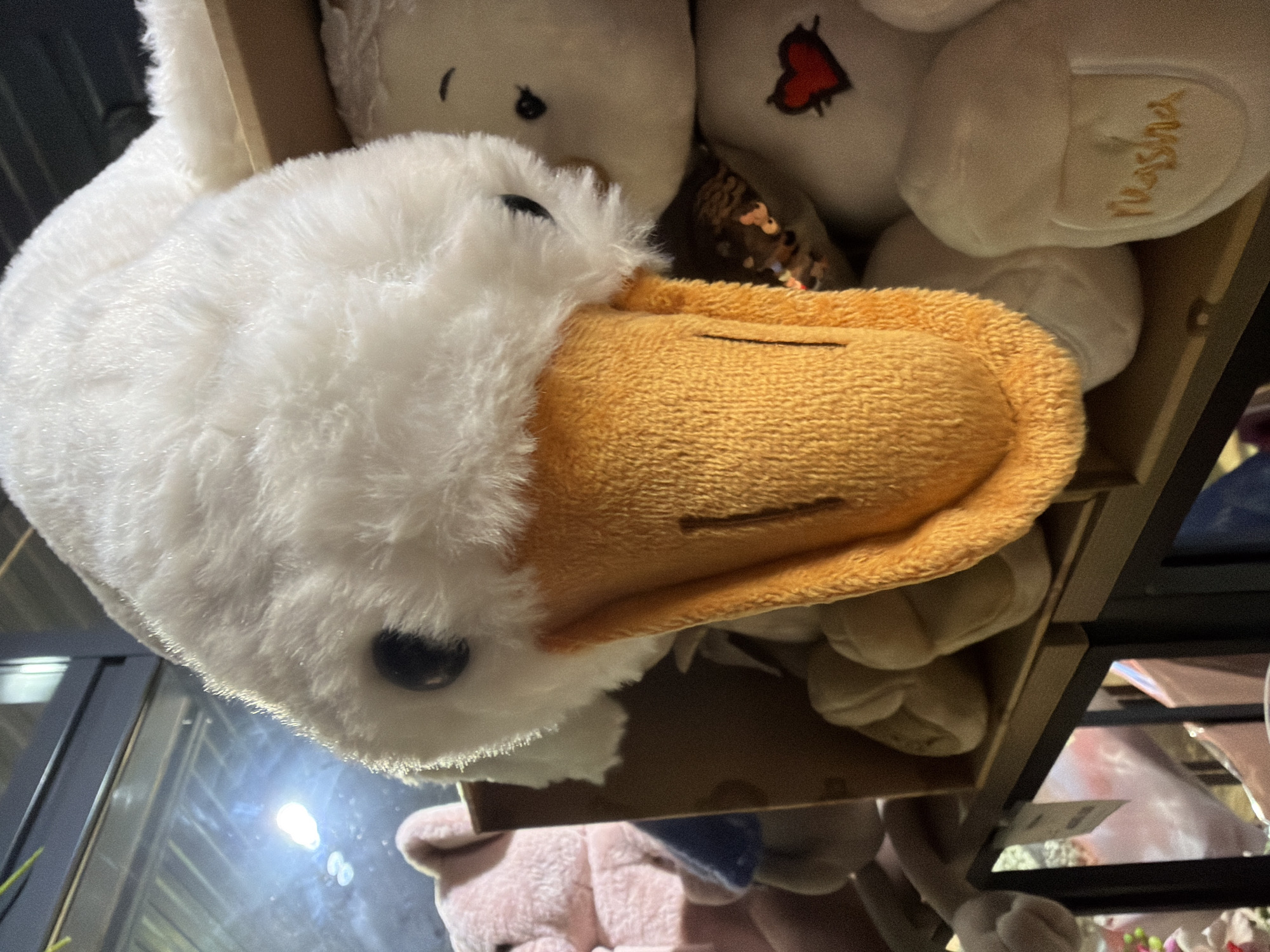 The image shows a plush white duck toy with an orange beak. The toy is positioned in a display shelf in a shop. Other plush toys, including a white bear with a heart and the name "Nusha," are also visible on the shelf. Part of a window and some light are seen reflecting off its surface. The lower part of the image shows other stuffed toys in the shelf below. It is not possible to confirm that the image was taken in Chaikovsky, Russia.