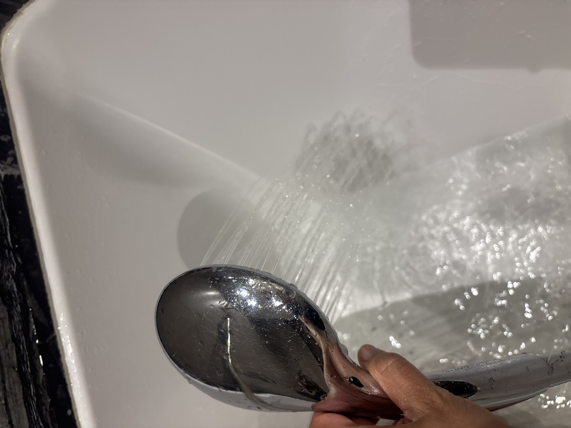 A hand is holding a chrome shower head, from which water is spraying into a white bathtub. The shower head is positioned in the upper left quadrant of the image, and the spray is directed diagonally down and to the right, creating a misty effect. The bathtub's surface is white and appears to be made of porcelain or a similar material. The edges of the bathtub are visible, with a dark, possibly granite, countertop or surround along the top edge. The scene is likely taking place in a bathroom. The image does not provide any specific visual cues to determine its location within Dubai, United Arab Emirates.