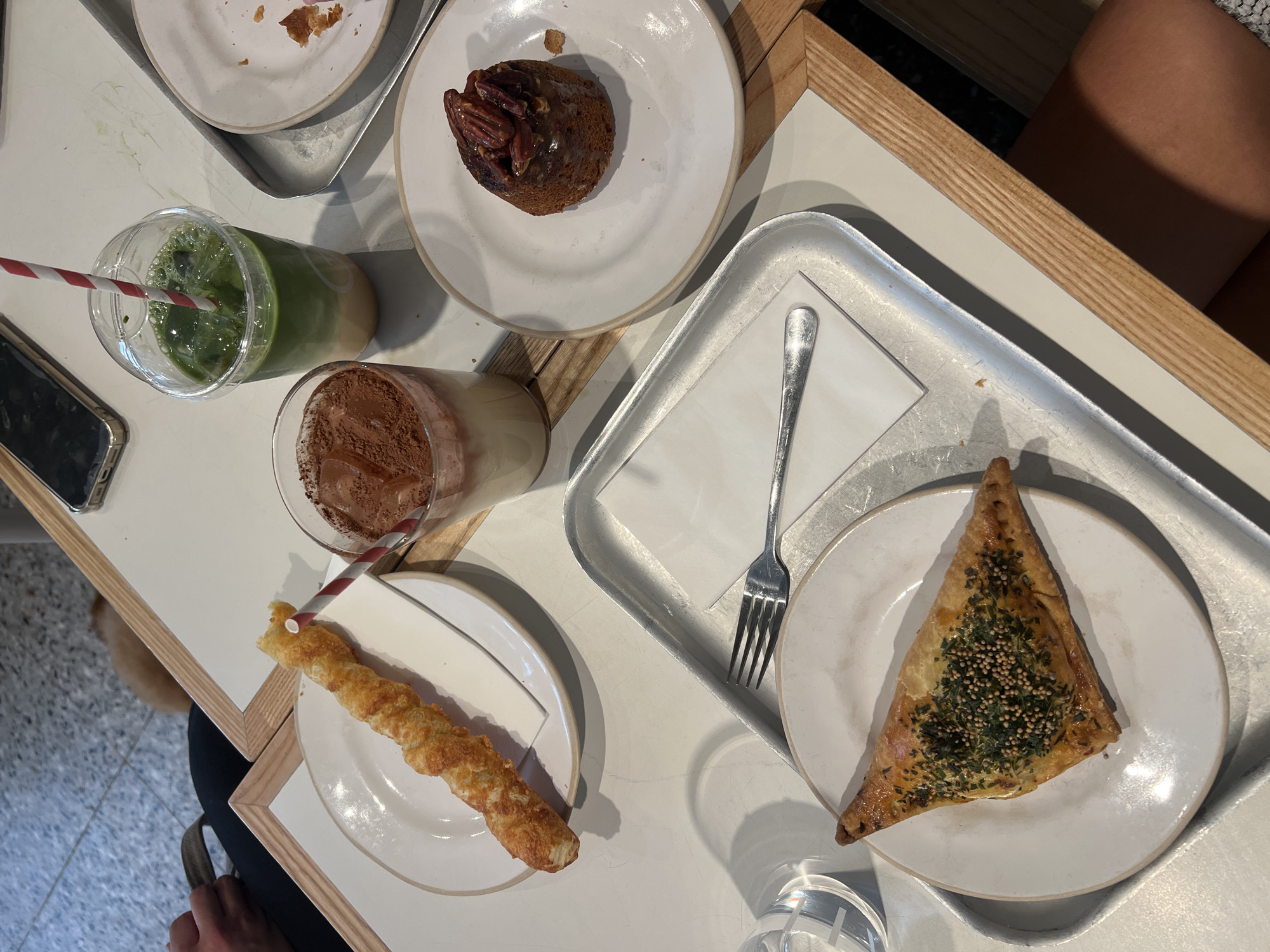 The image shows a close-up, high-angle view of a table at a cafe, likely in Manchester, United Kingdom, with various food and drink items. In the foreground, on a white plate, is a triangular pastry topped with herbs and seeds. To the left, a glass of water is visible. Above this, on a metal tray, is a fork resting on a white napkin. Further up and to the right, on another white plate, is a small, round cake topped with pecans and drizzled with caramel. Beside it, on a white tray, is a plastic cup containing a green beverage, possibly matcha, with a red and white striped straw. To the left of the green drink, a glass holds a beverage with a foamy top dusted with cocoa powder and ice. Next to that, on a white plate, is a twisted breadstick with cheese. A smartphone is visible on the table in the background. The table itself is white with wooden trim. A person's arm and shoulder are visible in the lower right corner, and another person's hand and arm are seen in the upper left corner, suggesting a social setting. The floor beneath the table appears to be a speckled tile.