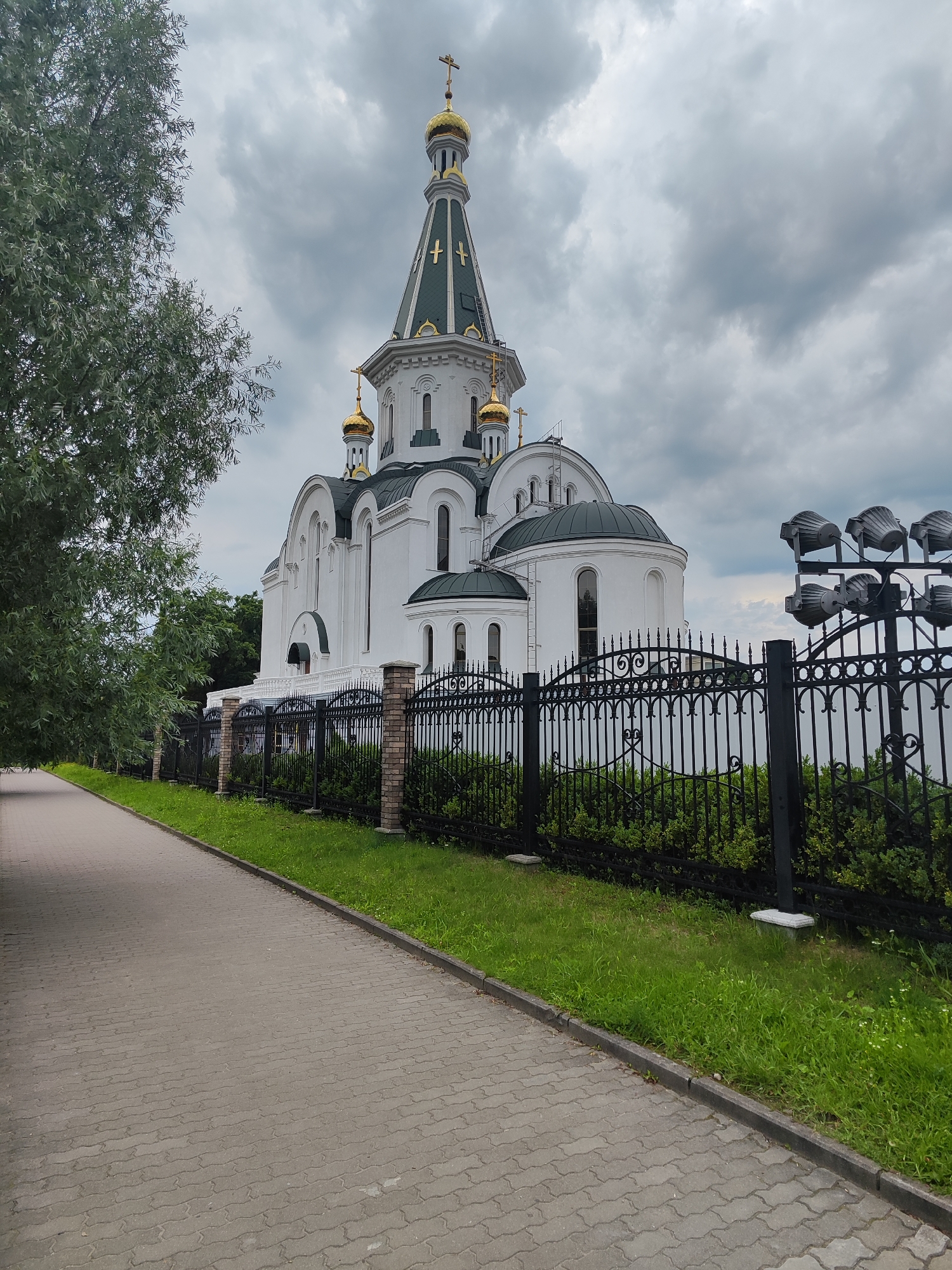 The image shows an Orthodox church in Kaliningrad, Russia, under a cloudy sky. The church is white with green roofs and golden domes topped with crosses. A black metal fence with brick pillars encloses the church grounds, separating it from a paved walkway that runs along the left side of the image. Lush green grass and bushes are visible behind the fence. To the right of the church, a row of outdoor spotlights is mounted on a pole. A large tree with green foliage is situated on the left side of the walkway, partially obscuring the view of the sky and the upper portion of the church. The scene appears to be captured during the daytime.