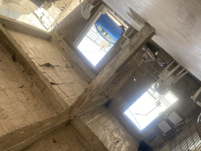 The image depicts an interior space within a concrete structure located in Street, Nigeria. The floor is tiled with light-colored square tiles. The ceiling consists of exposed concrete beams and slabs, showing visible cracks and exposed reinforcing bars in various sections. Walls are constructed from concrete blocks.
Two rectangular openings provide natural light and views to the exterior. Through the upper-left opening, bright sky is visible, along with a street light pole and a metal security gate. Below this opening, on the interior wall, three white box-shaped electrical units are mounted, accompanied by electrical conduits. A stack of three white plastic chairs is positioned beneath these units, alongside a collection of unidentifiable items.
The mid-right opening reveals a portion of a blue sedan parked outside the structure. A single white plastic chair is situated in the lower-right area of the frame. No individuals are present in the scene. The area is brightly illuminated by daylight entering through the openings.