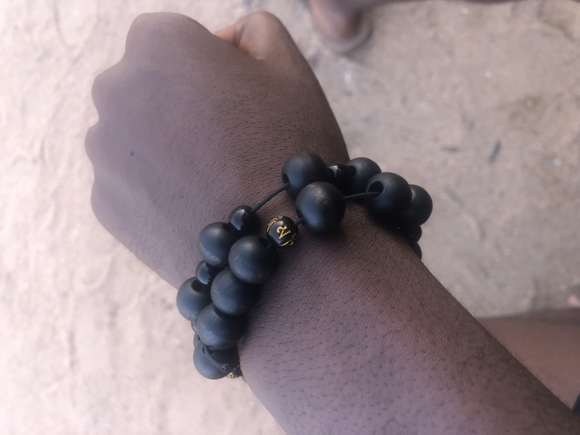A close-up shot shows a person's left forearm and hand, adorned with a multi-strand bracelet composed of black, matte wooden beads and a few smaller, shiny black beads. One of the shiny beads is accented with a small gold charm featuring an emblem. The bracelet is wrapped around the wrist and forearm. The skin tone is dark brown, and the background consists of a textured, light brown surface, possibly sand or dry earth, suggesting an outdoor setting. The image appears to be taken in Jalingo, Nigeria, as indicated by the prompt. The focus is on the bracelet and the texture of the skin, with the background blurred.