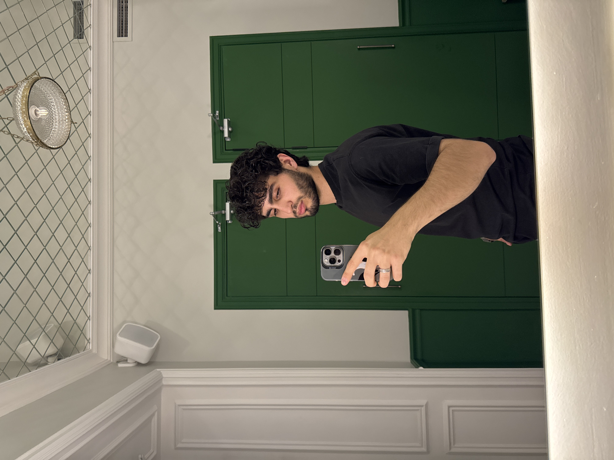 A man with dark curly hair and a beard is taking a picture of himself in a mirror. He is holding an iPhone in his right hand. The background includes a green wall cabinet, a white tiled wall with a decorative light fixture, and white wainscoting. The location appears to be a bathroom. There is no information available in the image that specifies the location as being in Yerevan, Armenia.
