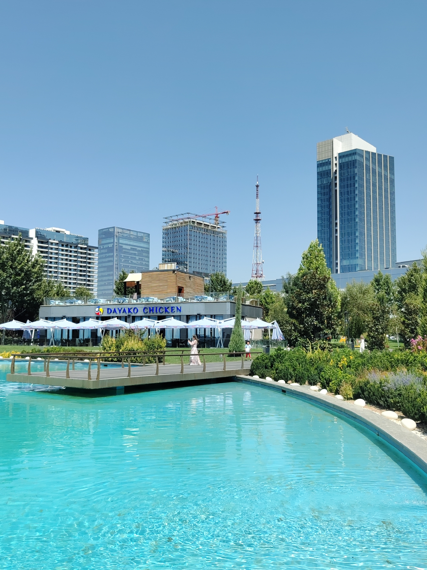 This is a photograph taken in Tashkent, Uzbekistan, at the "Dayako Chicken" restaurant and its surrounding area. In the foreground, a bright blue, shallow pool of water is visible, with a wooden walkway extending into it. The walkway has metal railings.
In the middle ground, there is a restaurant with an outdoor seating area covered by numerous white umbrellas. The restaurant's signage, "DAYAKO CHICKEN," is clearly visible in blue lettering on the building. A woman in a light-colored dress is standing on the wooden walkway, appearing to take a photo with her phone. Several other people are visible in the background, near the restaurant.
Behind the restaurant, several modern high-rise buildings dominate the skyline. These include a tall, glass-faced skyscraper on the right, and other multi-story buildings to the left and center. A construction crane is visible on one of the buildings. A distinctive telecommunications tower with red and white banding also stands tall in the background.
The environment is characterized by lush green trees and well-maintained landscaping surrounding the pool and restaurant. The sky is clear and blue, indicating a sunny day. The overall scene depicts a modern urban area with a recreational and dining space.