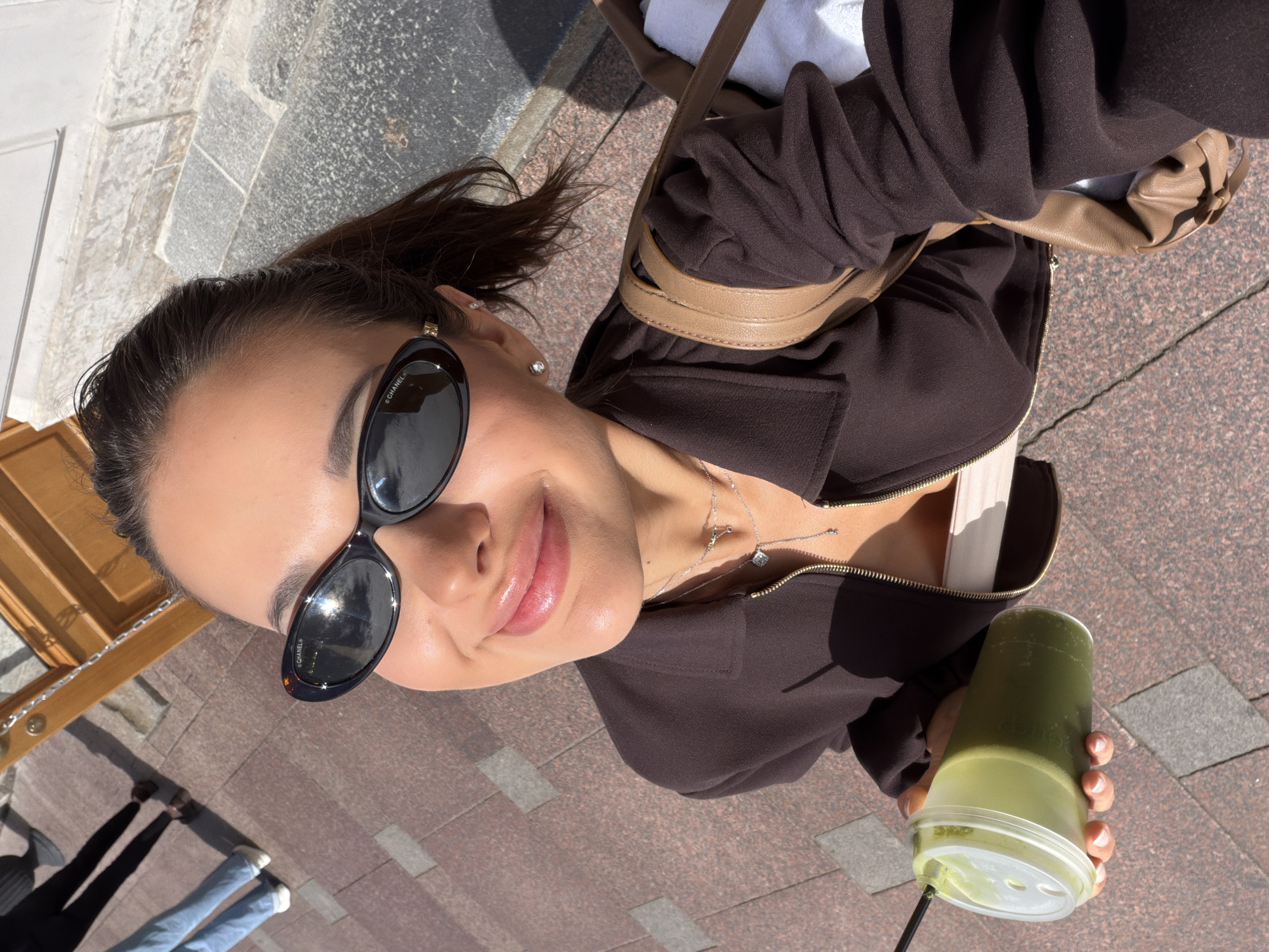 An overhead, high-angle selfie shows a young woman holding a green beverage in a clear plastic cup with a straw. She is wearing sunglasses, a brown zip-up jacket, and a light-colored top underneath. She is also wearing jewelry, including earrings and necklaces. A brown handbag is visible over her shoulder. The background consists of a cobblestone street with some shadows. The woman is smiling at the camera. The scene appears to be outdoors in Saint Petersburg, Russia, likely during daylight hours, given the bright sunlight and shadows. The cobblestone pattern is characteristic of many historic streets in Russian cities.