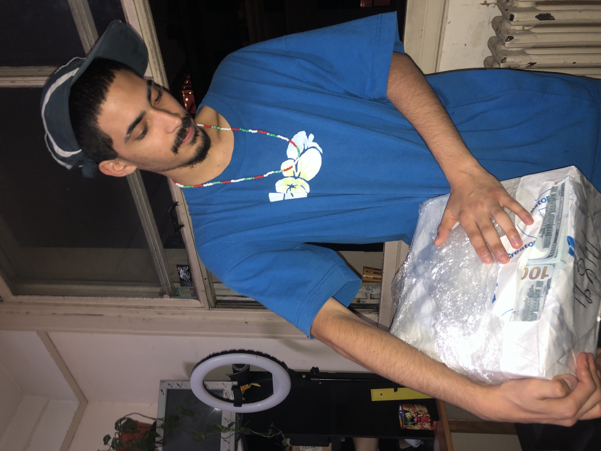 A man is holding a package wrapped in clear plastic and white paper with $100 bill imagery. He is wearing a blue t-shirt with a white floral design and a cap. He also has a beaded necklace. The background shows a window with stickers and what appears to be a radiator, a plant, a ring light, and a TV. The location is possibly indoors. The image does not provide information about its location in the city of Buenos Aires, Argentina.