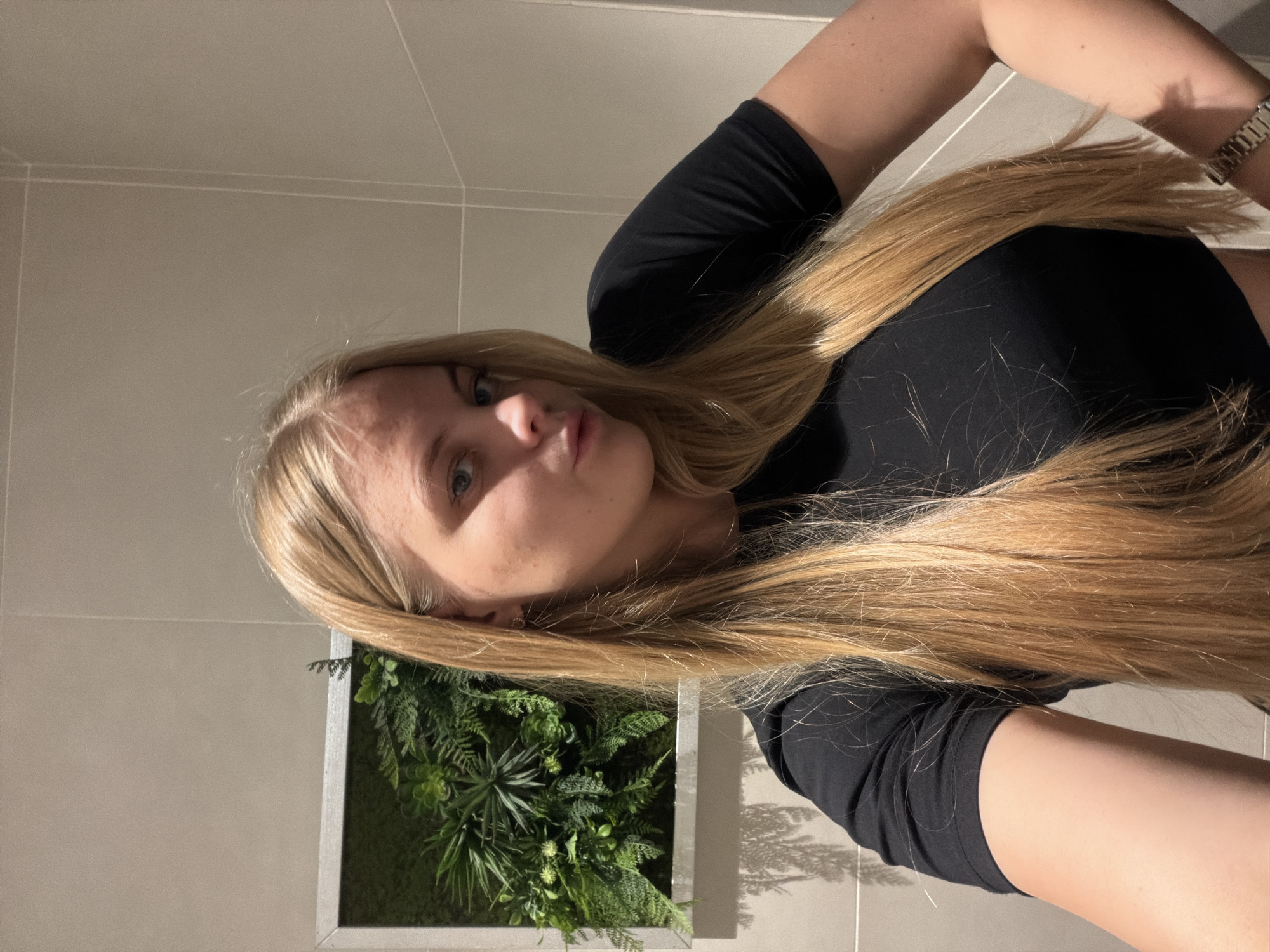 A young woman with long blonde hair is facing the camera in what appears to be a bathroom setting. She is wearing a black shirt. A wall decoration of green plants in a silver frame is visible behind her. Light gray tiles cover the wall. A small portion of a silver wristwatch is visible on her right wrist. The photo is taken indoors under artificial lighting. The location is not discernible as Izhevsk, Russia from the given image.