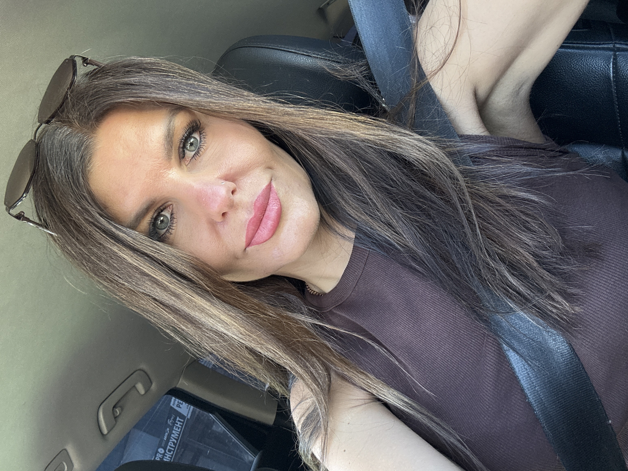 A woman with long, brown hair is taking a selfie in a car. She is wearing a brown tank top and has sunglasses on her head. A seatbelt is visible across her chest. The background shows the interior of the car, including the dashboard and window. The text on the window is in Cyrillic and appears to be advertisements for tools. The overall scene is consistent with a person taking a photo while sitting in the driver's seat of a vehicle in an urban or suburban environment. No specific location in Chaikovsky, Russia is identifiable from the image.