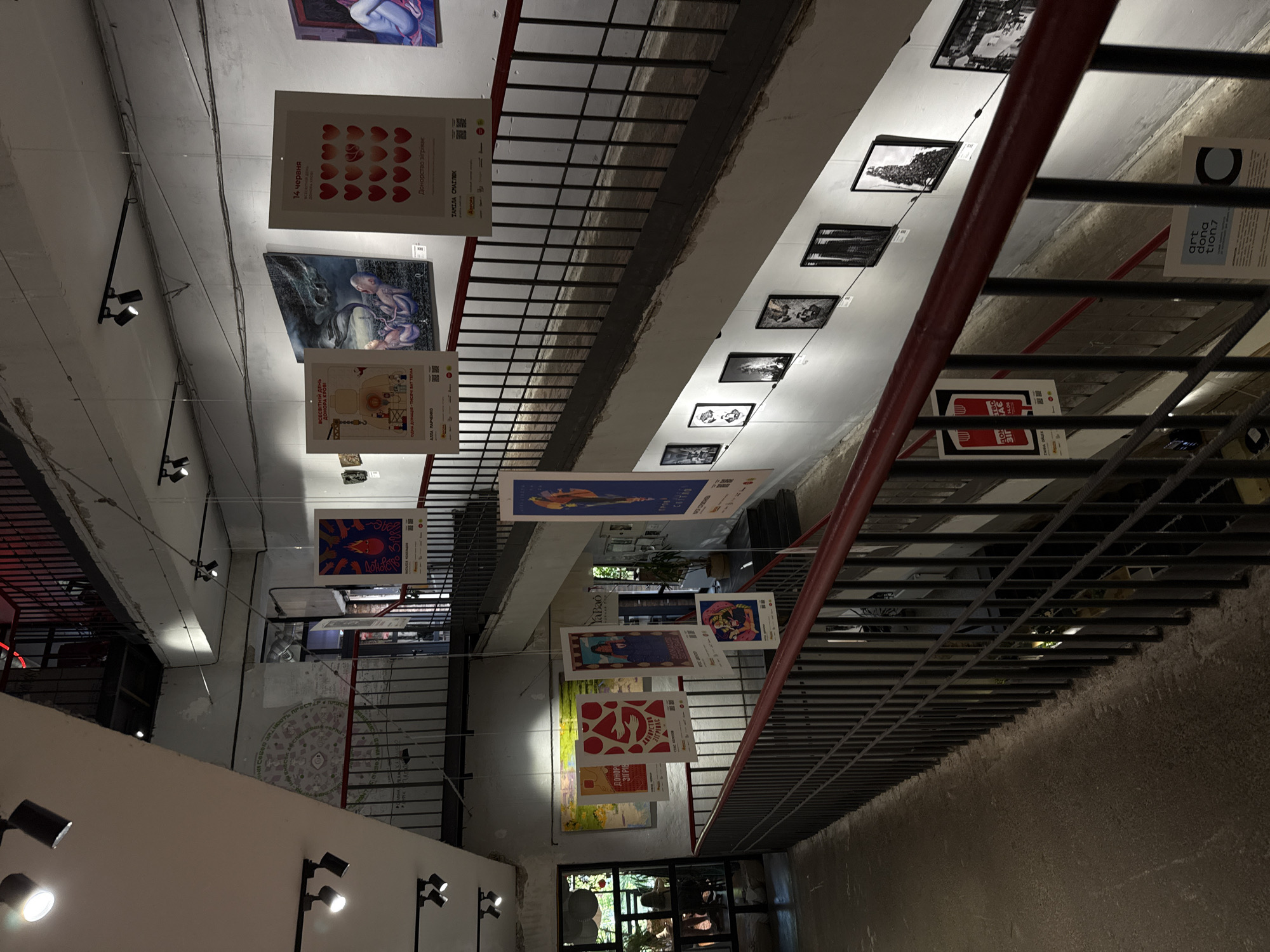 This is a photograph of the interior of a multi-level art gallery or exhibition space. The scene appears to be in Kyiv, Ukraine, suggested by the Cyrillic text on the posters. The architectural style is industrial, with exposed concrete walls and ceilings, and metal railings for the staircases and walkways. Spotlights are mounted on the ceiling, illuminating the exhibition.
On the walls, various pieces of art are displayed, including posters and framed artworks. Several posters, predominantly in red, white, and yellow, are suspended from the ceiling at different levels. These posters contain text in Cyrillic and images, some depicting abstract designs and human figures. One poster features a series of red hearts. Another, to the left, shows a stylized illustration of hands holding a heart with flames. Further down the wall, a larger artwork depicts a surreal scene with two infants and a creature emerging from smoke. Black and white framed photographs are displayed on the lower wall sections, arranged in a gallery style.
The space has a mezzanine level with a balcony overlooking the lower section, connected by a staircase with a dark metal railing and a red handrail. The floor in the foreground appears to be concrete, with some texture. In the background, through glass doors, a glimpse of an outdoor area with greenery and patrons can be seen. One person is visible standing near the glass doors on the left. The overall atmosphere is that of a contemporary art exhibition venue.