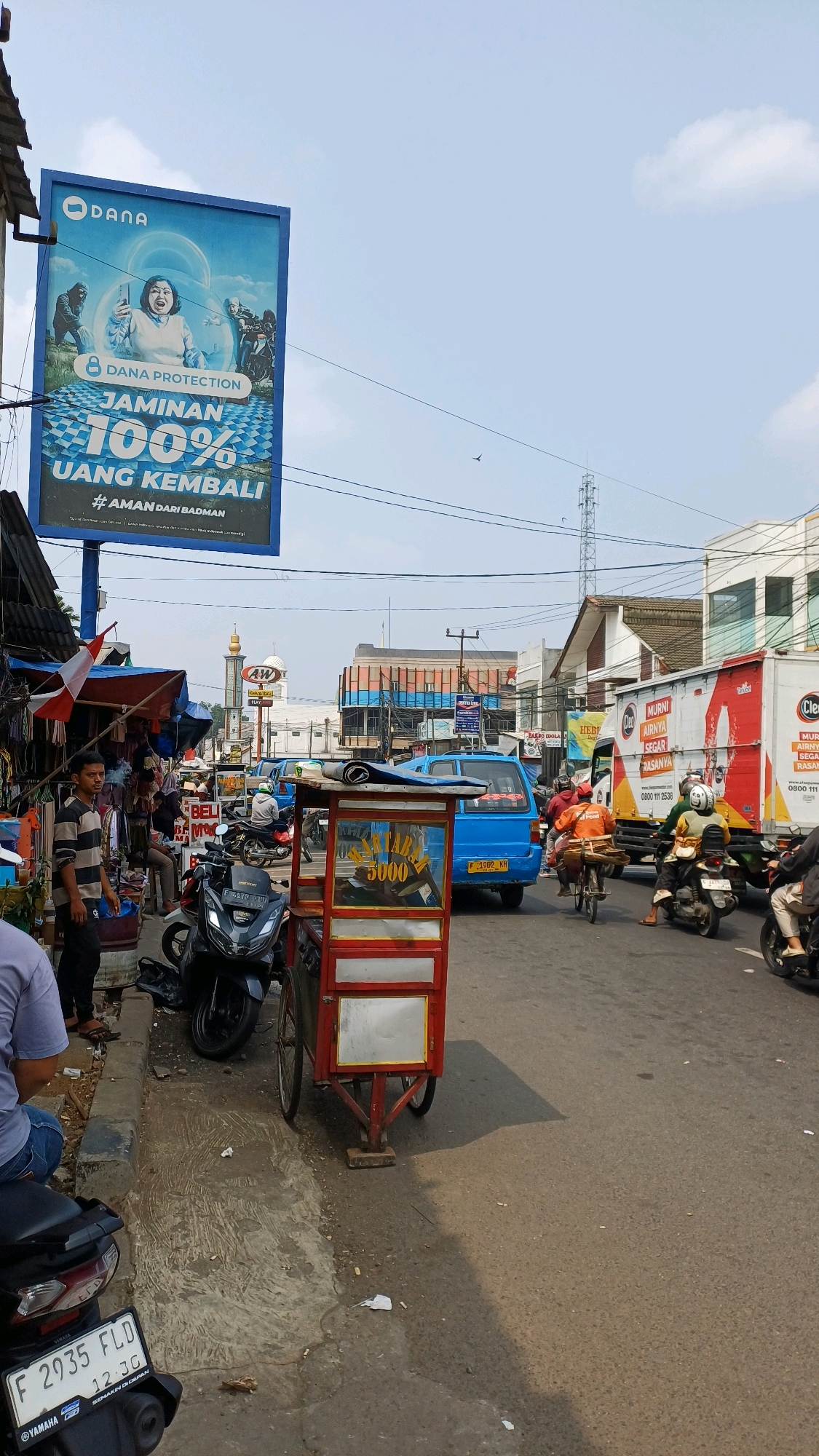 The image depicts a street scene in Citeureup, Indonesia. A large blue billboard for "DANA PROTECTION" with promotional text in Indonesian advertising "JAMINAN 100% UANG KEMBALI" is prominently displayed on the left. Below the billboard, a market stall with various goods, including clothing and what appears to be flags, is set up along the sidewalk. Several motorcycles are parked on the side of the road. A red and yellow food cart is positioned on the street. The road is busy with traffic, including a blue van, several motorcycles with riders, and a white truck with "HEBAT" and "URUS" text. People are visible in the scene, including a man standing near the market stall, a person sitting on a motorcycle, and riders on other vehicles. In the background, a mosque with a minaret and an "AW" restaurant sign are visible, along with other buildings and a cell tower. The sky is partly cloudy. The overall impression is of a bustling commercial area on a sunny day.