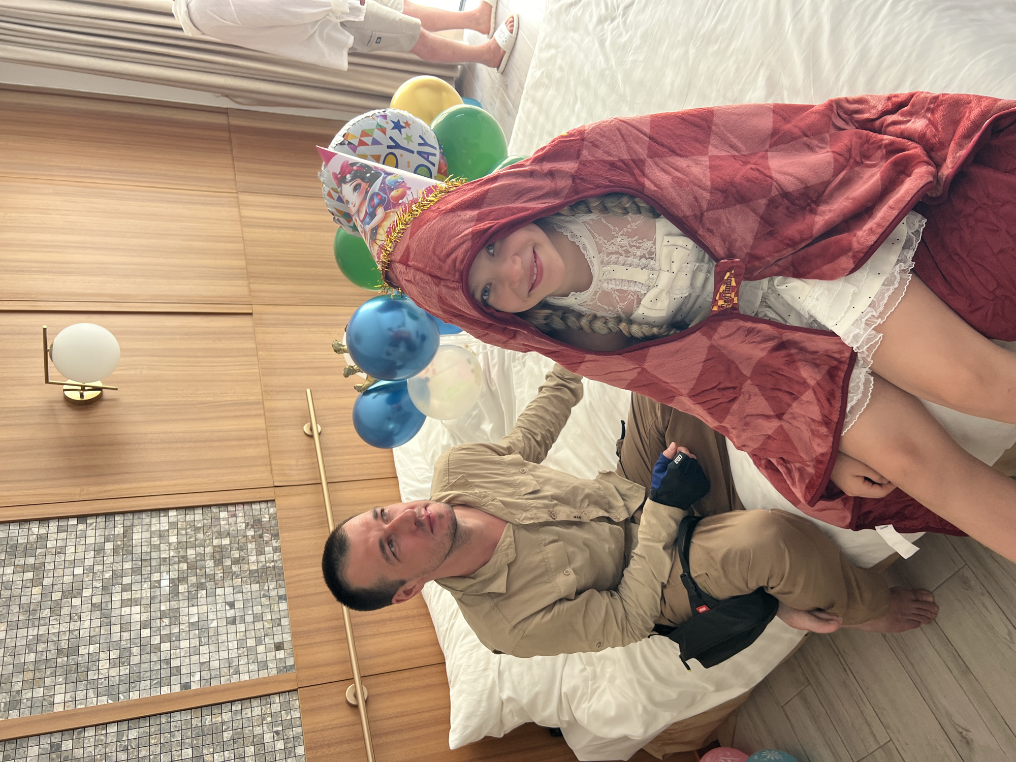 In this media file, two people, a young girl and an adult male, are the subjects. The girl is lying down, wrapped in a red blanket that appears to have a checkered pattern, and she is wearing a white lace dress and a birthday hat with a cartoon character on it. She has blonde hair braided into pigtails and is smiling. The adult male is also lying down next to her, wearing a tan button-down shirt and brown pants. He has short dark hair and appears to be looking at the girl.
Surrounding them are several balloons, some of which are blue, green, and yellow. One balloon has a cartoon image of Snow White on it, matching the girl's hat. The scene appears to be indoors, possibly on a bed covered with white bedding. The background consists of wooden paneled walls and a section of tiled wall. There is also a light fixture on the wall.
The presence of balloons and a birthday hat suggests that a birthday celebration is taking place. The overall mood seems cheerful and celebratory, with the girl's smile indicating happiness. There is no discernible time of day or weather information from the image. There is faint text on the man's shirt that appears to read "MAN".