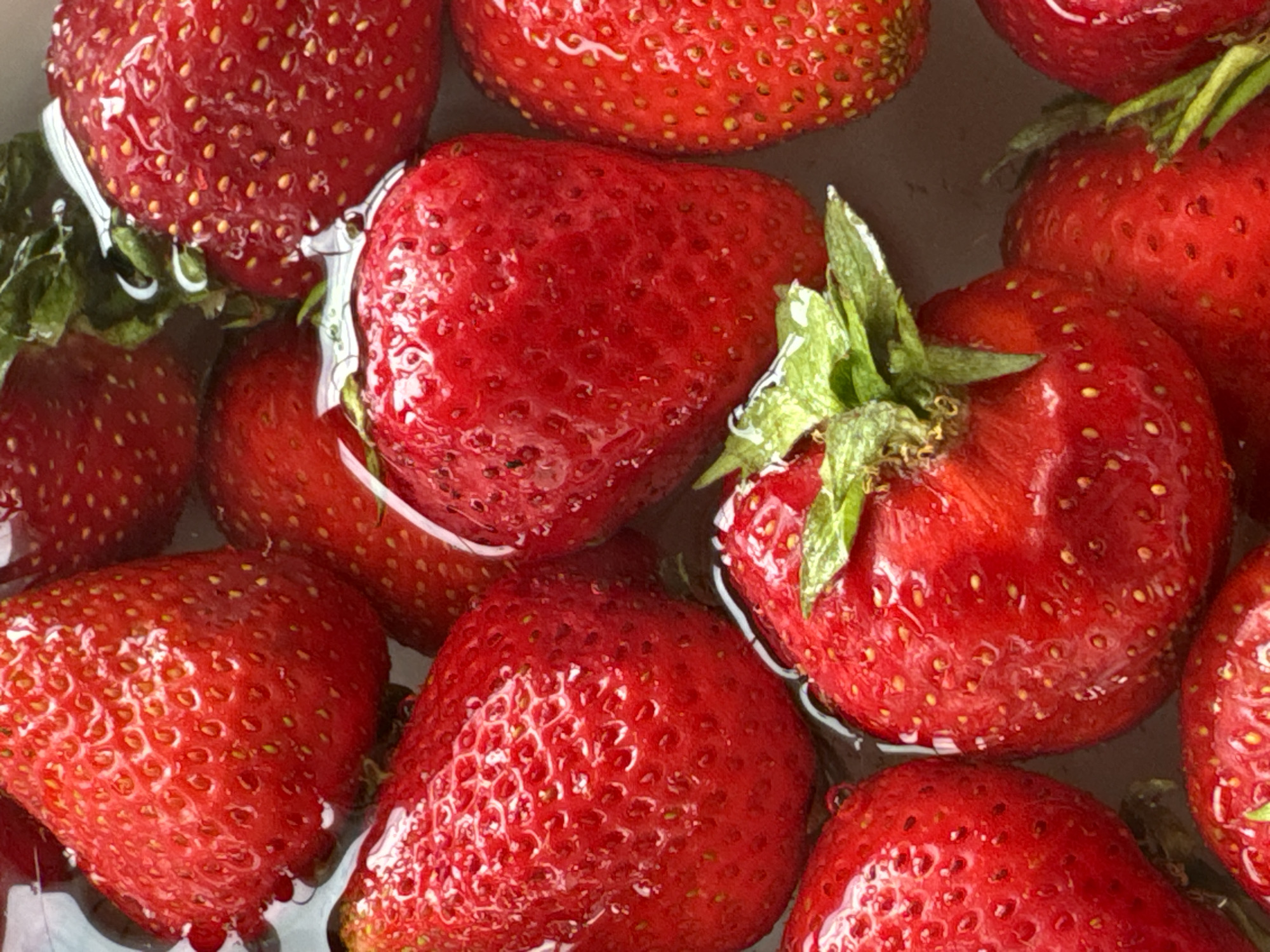 The image is a close-up, overhead shot of several ripe strawberries submerged in water. The strawberries are a vibrant red and have visible seeds on their surface. Some of the strawberries have green leaves attached at the stem. The water is clear, and reflections are visible on the surface of the strawberries. The scene appears to be a bowl or container filled with water and strawberries, possibly for washing or preparation. There is no indication of a specific location in Moscow, Russia, or any identifiable landmarks. The focus is solely on the fruit.