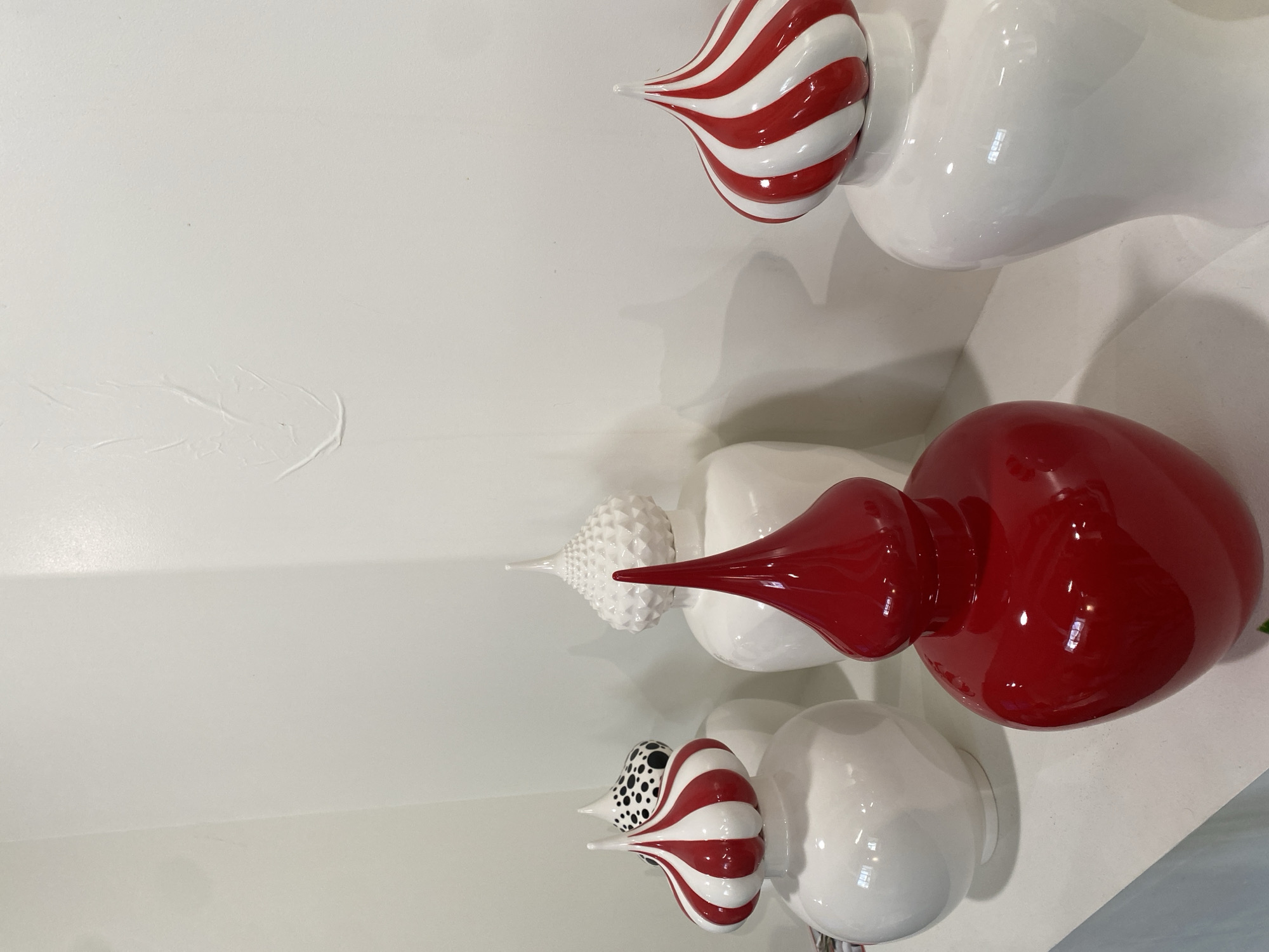 The image displays a collection of decorative, possibly ceramic, art objects mounted on a white wall. The objects feature bulbous shapes, some with striped patterns in red and white, while others are plain white, dotted black and white, or solid red. These objects appear to be stylized representations, potentially reminiscent of ornaments or decorative tops. The environment is a simple, indoor setting with a white wall. There is no indication that the image was taken in Moscow, Russia.