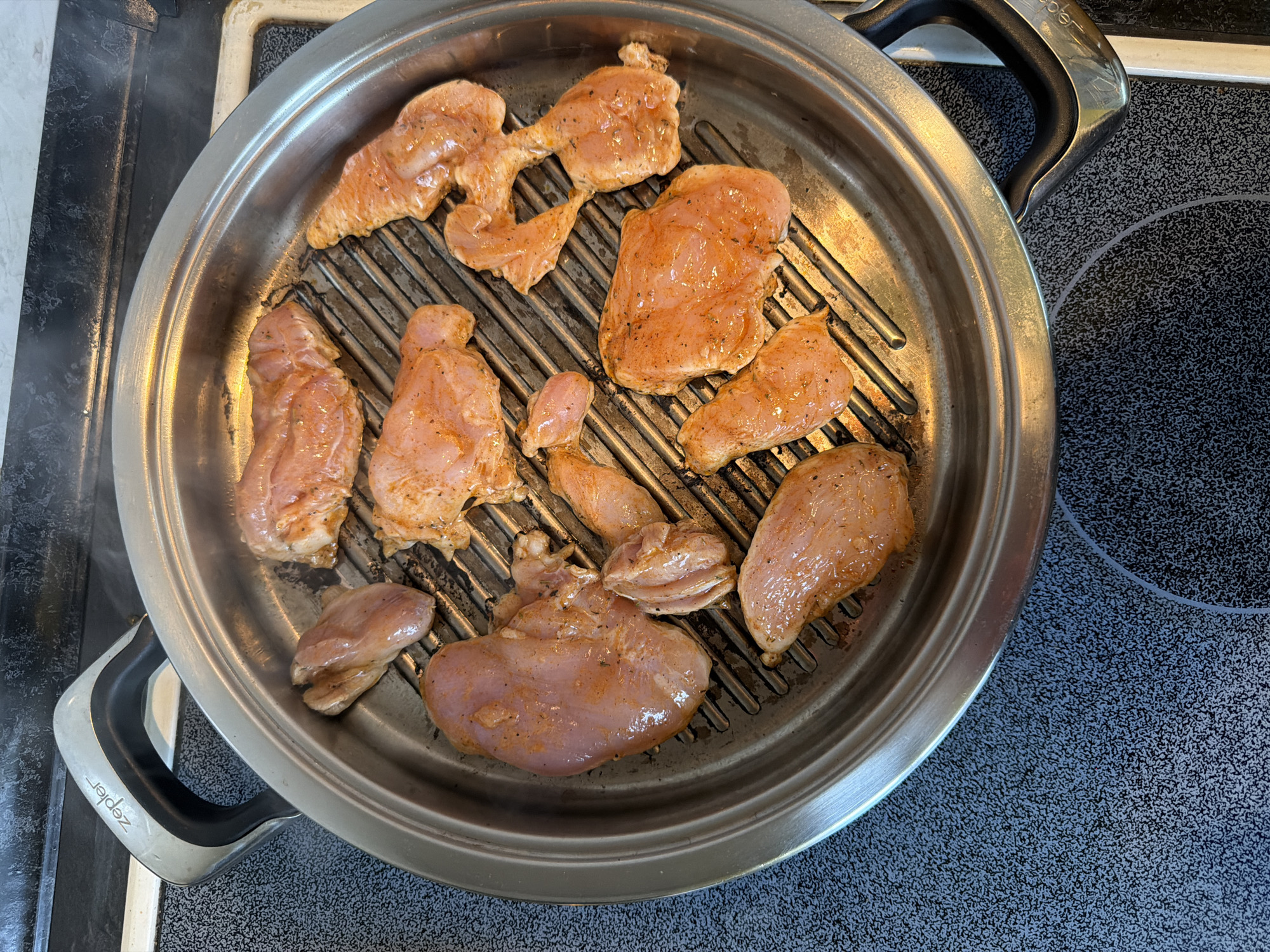 A high-angle shot depicts a silver-colored pan on a black stovetop. The pan contains several pieces of raw, seasoned chicken. Steam rises from the pan. The pan has two handles labeled "Zepler". The stovetop has a speckled gray and black pattern. The location and city of Pleven, Bulgaria, are not discernible from the visual information provided.