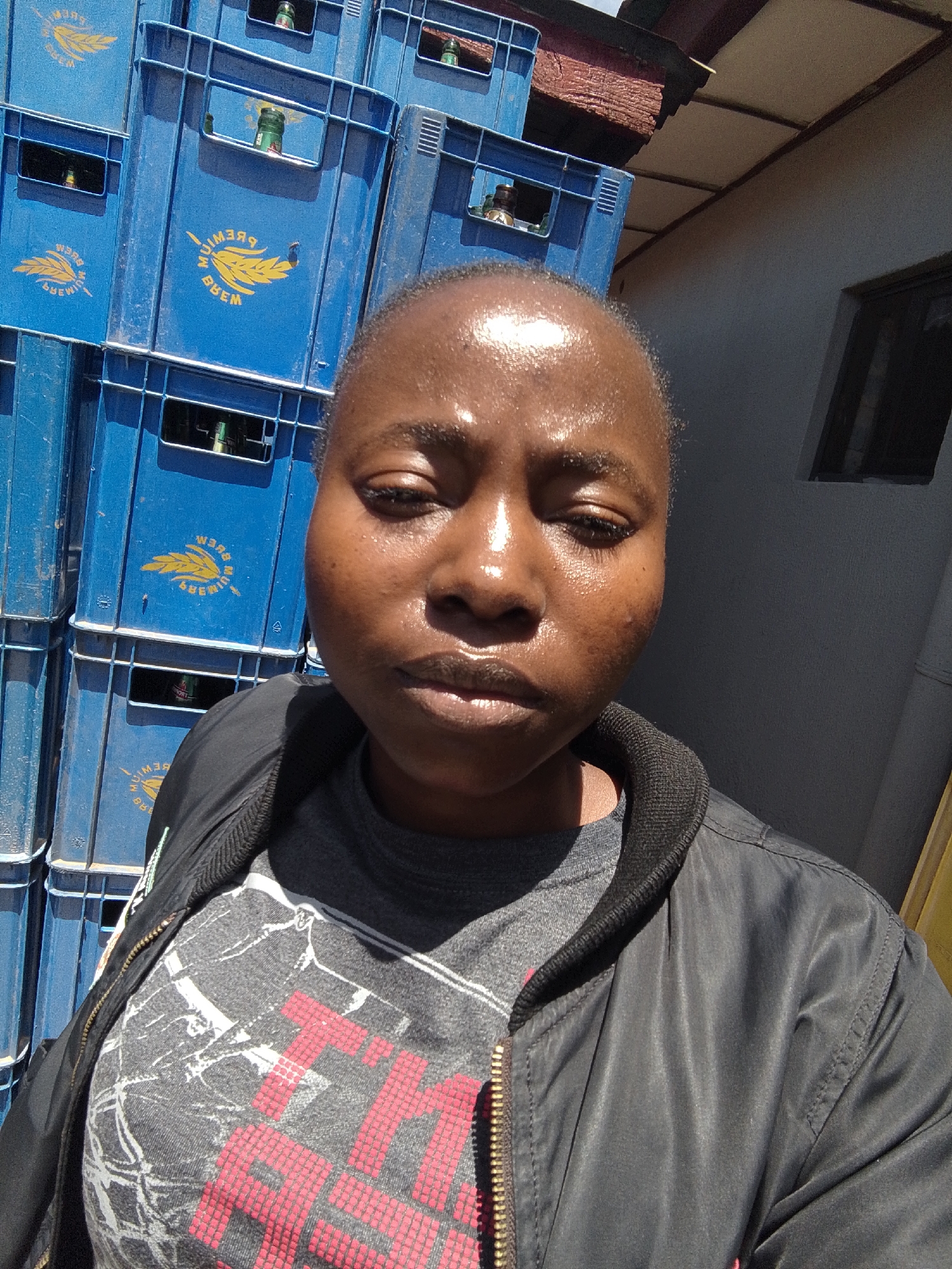 A woman is captured in a close-up, waist-up selfie taken outdoors. She is wearing a black bomber jacket zipped over a gray t-shirt with a graphic print. The background consists of stacked blue plastic crates, some of which contain bottles. The crates appear to be for beverage distribution. The lighting suggests it is daytime, with sunlight glinting off the woman's forehead and the surface of the crates. The scene is consistent with an outdoor area in a city like Jos, Nigeria, likely a commercial or storage space for drinks.