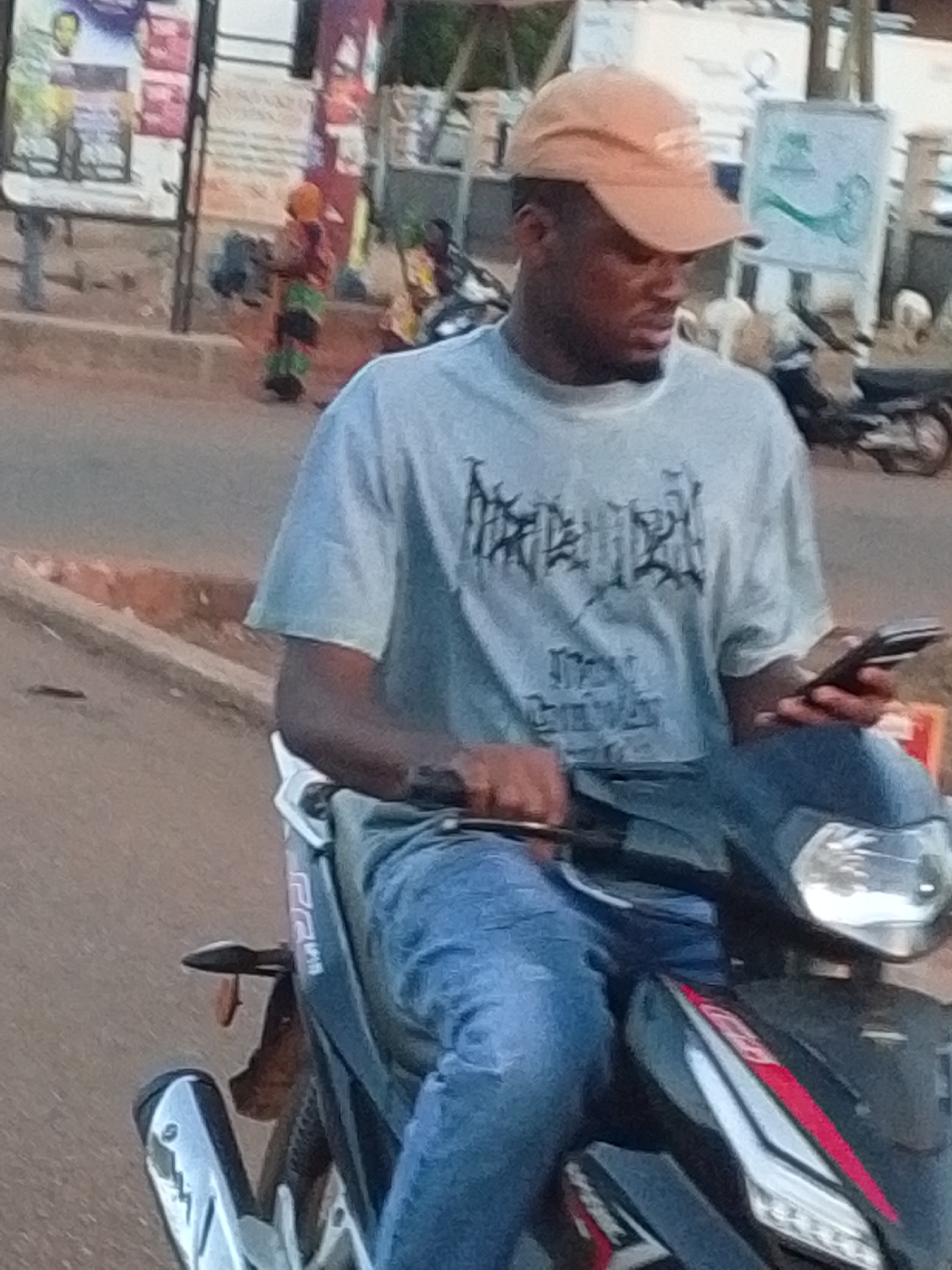 A man is riding a black and red scooter in Tamale, Ghana. He is wearing a light gray t-shirt with a black design, blue jeans, and a light brown baseball cap. He is looking down at a cell phone he is holding in his left hand while steering the scooter with his right hand. The road is paved and the background includes sidewalks, parked motorcycles, buildings, and a sign with green writing on it. There is also a woman with luggage standing on the sidewalk in the background.