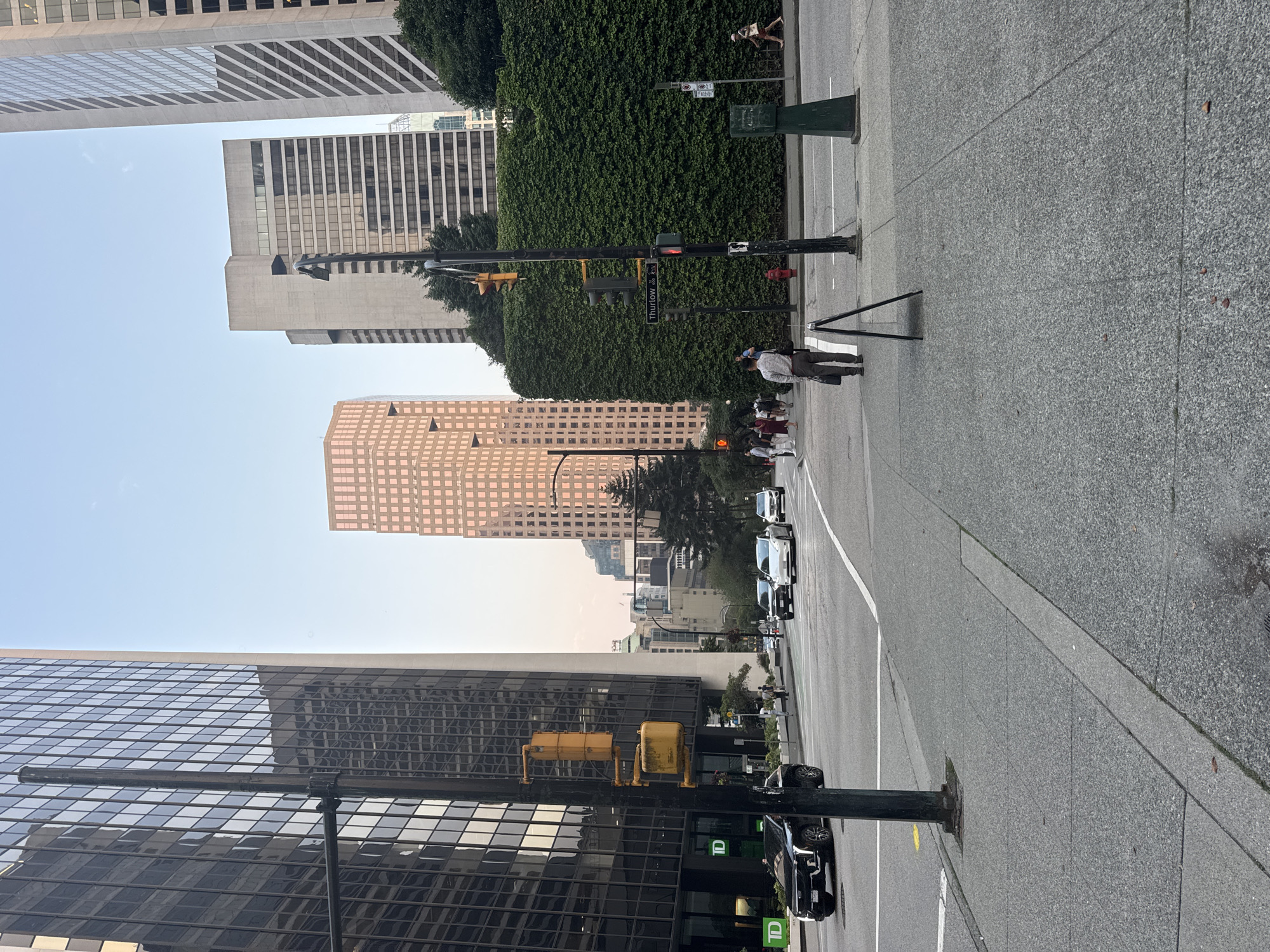 This image depicts a street scene in Vancouver, Canada, likely in the downtown core, characterized by a mix of modern skyscrapers and urban infrastructure. The perspective is from a sidewalk looking towards a street intersection with several vehicles and pedestrians.
Dominating the background are several tall buildings. On the left, a dark-glass skyscraper with a grid-like facade rises prominently. To the right of this, a lighter-colored, modern building with a textured facade is visible. Further in the background, a distinctive pinkish-toned hexagonal building stands out. The sky above is a clear, pale blue, suggesting it is daytime, likely late afternoon or early evening given the soft lighting.
In the mid-ground, the street is moderately busy. Several cars are visible, including a black SUV on the left, a white sedan in the center lane, and other vehicles further down the road. Traffic lights are present at the intersection. A large, well-maintained green hedge forms a prominent feature on the right side of the street, obscuring a building behind it.
Pedestrians are scattered throughout the scene. On the right sidewalk, a man in a light-colored shirt and dark pants is walking away from the camera, carrying a dark bag. Other pedestrians are visible near the crosswalk, some waiting to cross.
On the left side of the street, a TD Bank branch is identifiable by its green signage on the dark-glass building. A traffic light pole with yellow signal heads stands in the foreground, positioned on the sidewalk. A temporary A-frame sign is placed on the sidewalk in front of the man in the light-colored shirt.
The ground surfaces consist of gray asphalt for the street and textured concrete for the sidewalks, with visible expansion joints and some scattered debris on the sidewalk. The overall impression is of a busy, well-ordered urban environment.