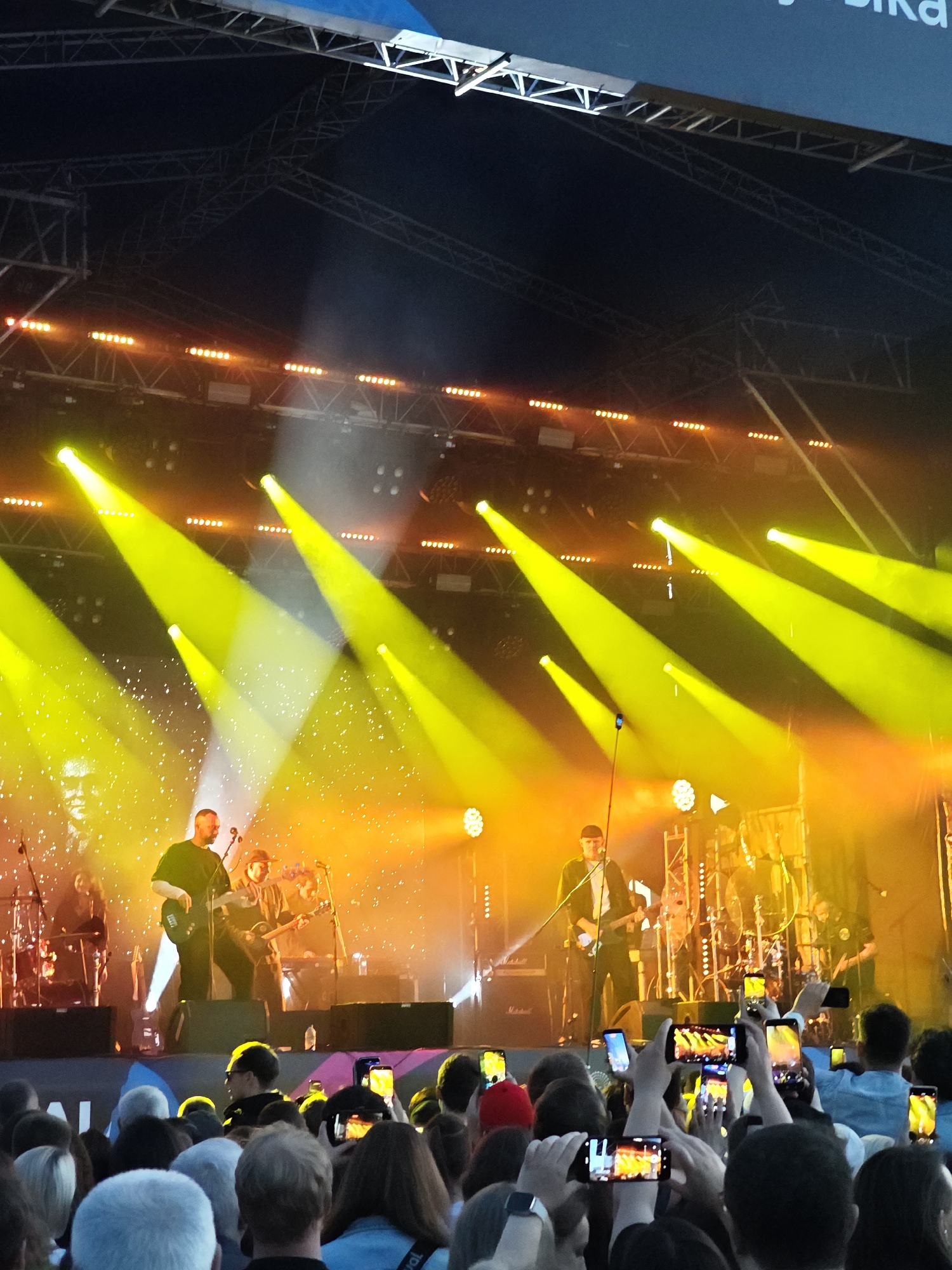 The image depicts an outdoor concert scene at night, likely at a venue within Yekaterinburg, Russia, given the context of the provided information. The stage is illuminated by numerous bright yellow spotlights and orange stage lights, creating a vibrant and dynamic atmosphere. On stage, several musicians are performing. In the foreground, a bald man in a dark t-shirt is playing a guitar. Behind him, another musician, wearing a cap, is also playing a guitar. Further back, a drummer can be seen behind a drum kit. The stage itself is equipped with speakers and lighting rigs.
In front of the stage, a large crowd of people is gathered, many of whom are holding up their mobile phones to record or photograph the performance. The audience members are diverse in appearance, and some are wearing casual clothing. The overall environment suggests a lively music event taking place in a public space within the city. The background shows stage structure and a large screen, possibly displaying visuals for the concert. The sky above is dark, indicating it is nighttime.