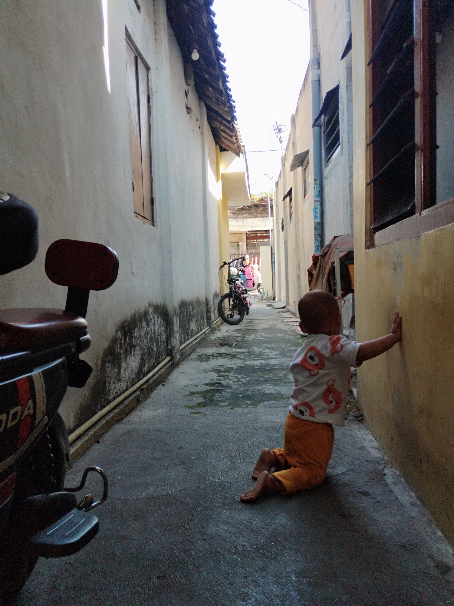 A young child, likely a toddler, kneels on a wet, paved alleyway, leaning their left hand against a cream-colored wall. The child wears a white t-shirt with a repeating pattern of orange cartoon bears and orange pants. The alley is narrow, with textured, light-colored walls on either side. A dark, slightly shiny motorcycle is parked on the left side of the alley, in the foreground. The motorcycle has a red seat and is partially visible. Further down the alley, a bicycle is leaning against a wall, and two people are visible on a scooter or motorcycle in the distance. Sunlight streams down from the right, illuminating parts of the alley and casting shadows. The environment appears to be a residential area in Surabaya, Indonesia, characterized by its narrow passageways and the presence of motorcycles and scooters.