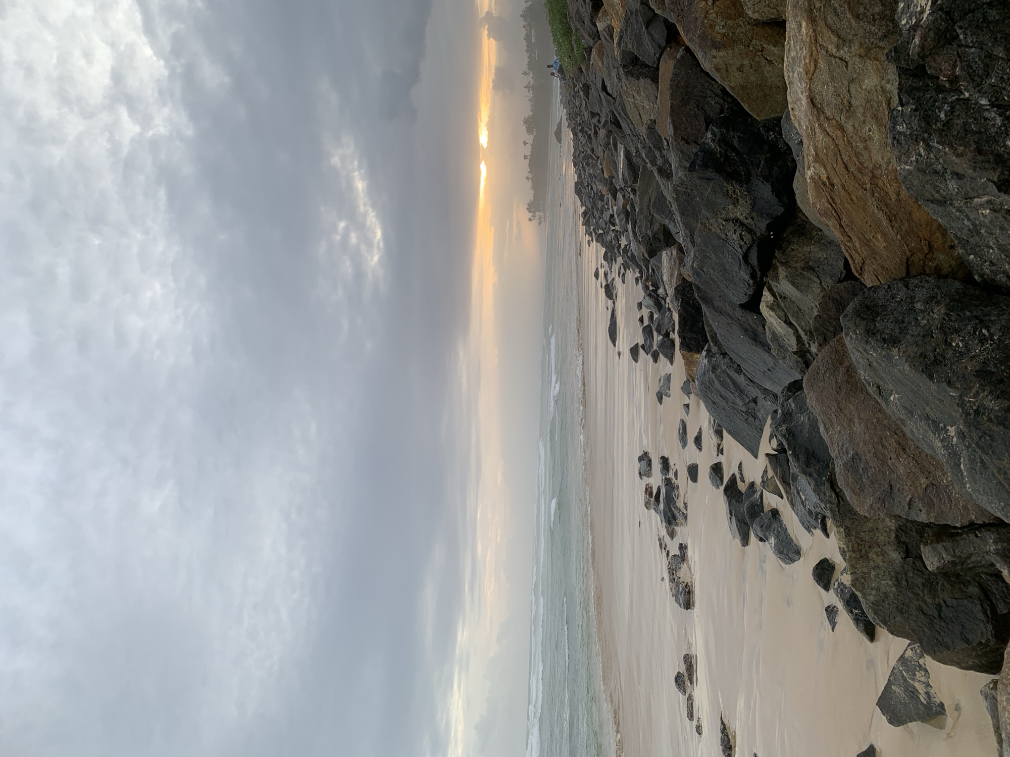 The image shows a beach in Kathaluwa, Sri Lanka, at sunset. The sky is filled with gray clouds, but a sliver of golden light is visible on the horizon. The beach is sandy and lined with rocks and boulders on the right side. Small waves are visible in the water. In the far distance, figures of people can be seen at the edge of the ocean.