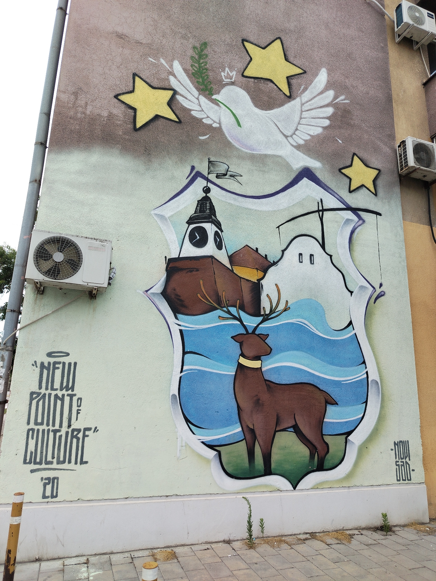 The image shows a mural on the side of a building, featuring a coat of arms with a brown deer in the foreground and a clock tower and other structures in the background, all within a blue, wavy sea. A white dove with a green olive branch in its beak flies above the coat of arms, accompanied by three yellow stars. To the left of the mural, the text "NEW POINT OF CULTURE '20" is spray-painted. To the right, the text "-NOV SAD-" is visible. Two white air conditioning units are mounted on the wall above and to the right of the mural. The mural is located in Novi Sad, Serbia. The ground in front of the building is paved with rectangular tiles, and a few small plants are growing through the cracks. There are also two bollards in the foreground.