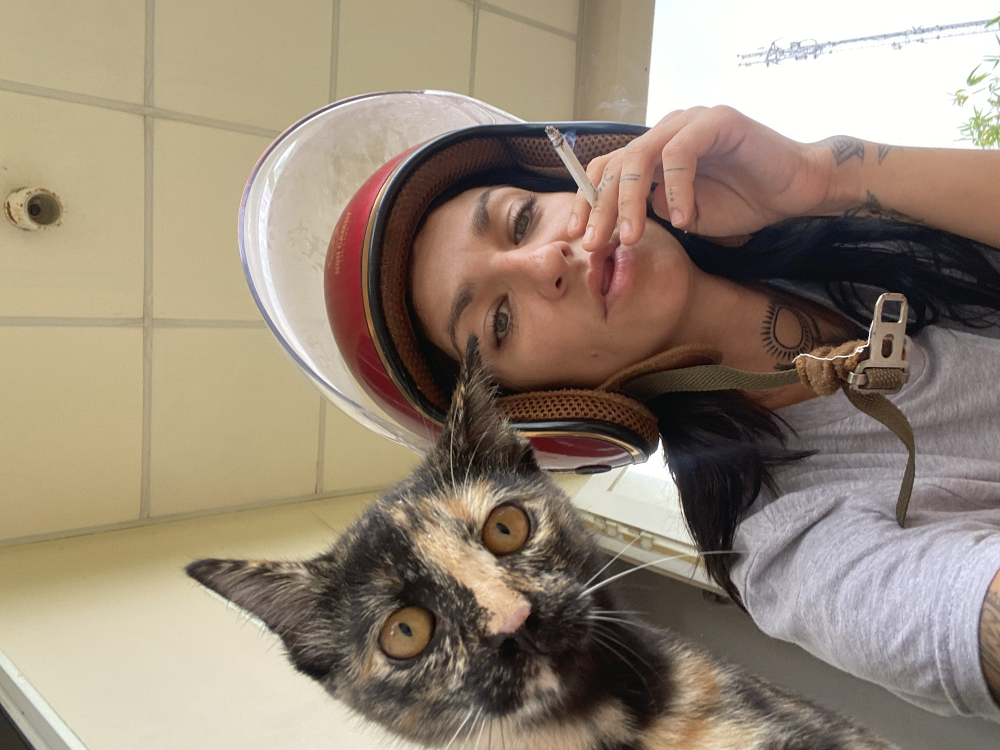 A low-angle, close-up photograph captures a woman and a cat in what appears to be a casual outdoor or semi-sheltered setting in Mũi Né, Vietnam, during the daytime. The perspective suggests a selfie or a shot taken from below.
The primary human subject is a woman with dark hair, looking directly towards the camera with a relaxed or slightly contemplative expression. She wears a red and clear motorcycle helmet, which is partly visible around her head. In her right hand, she holds a lit cigarette, with smoke subtly rising. Several tattoos are visible on her hand, arm, and upper chest, including a prominent sun-like design on her neck/collarbone area. She is wearing a light-colored t-shirt, and part of a khaki-colored helmet strap with a buckle is seen across her chest. The word "AVIATOR" is faintly visible on the side of her helmet.
In the foreground, a calico or tortoiseshell cat with distinctive black, orange, and cream fur patterns is looking intently at the camera. The cat's large, amber-colored eyes and prominent white whiskers are clearly visible, adding a curious and engaging element to the composition.
The background consists of light-colored, possibly tiled, walls or a ceiling, suggesting an architectural structure. Above the woman's head, an outdoor scene is visible with a bright, overcast sky, power lines, and some green foliage or trees. A circular, light-colored fixture is visible on the wall in the upper left of the frame. The overall lighting is bright and natural, indicating daylight. The image conveys a spontaneous and candid moment.