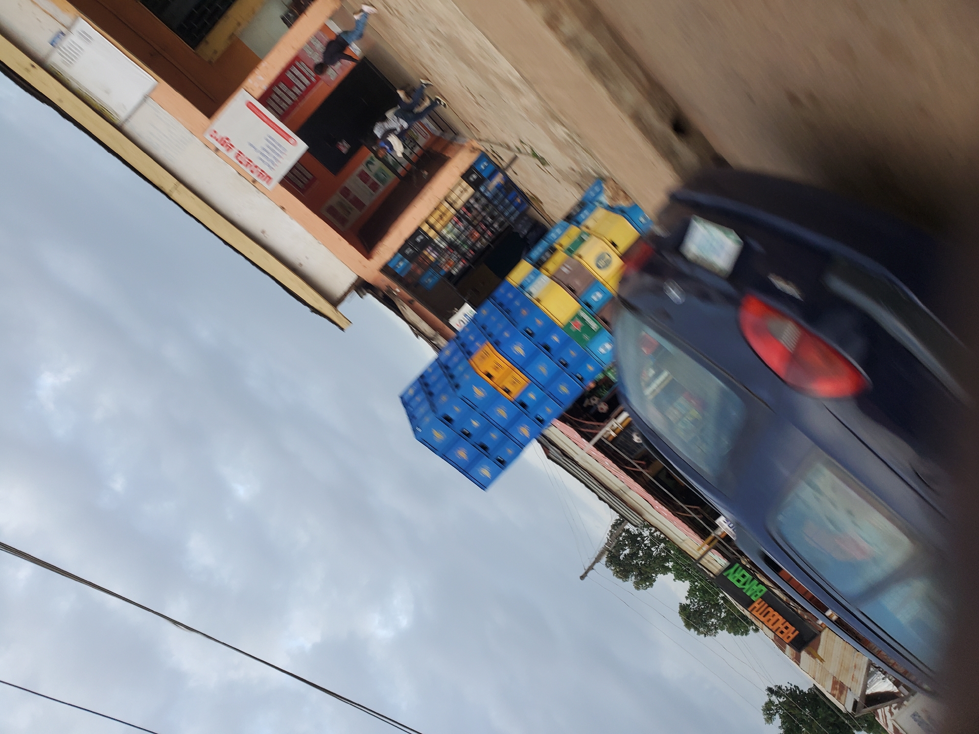 The image captures a street scene in Bukuru, Nigeria, on an overcast day. In the foreground, a dark-colored car is in motion, with its rear visible. To the left of the car, a sign reads "REHOBOTH BAKERY" in orange and green lettering. Behind the car, a building with an orange facade features an advertisement sign with red and white text. In front of the building, stacks of blue and yellow crates, likely containing beverages, are piled high. Several people are visible in front of the building, with one person walking away from the camera towards the right. The street is paved with dirt, and utility wires are visible overhead. The overall impression is one of a bustling commercial area.