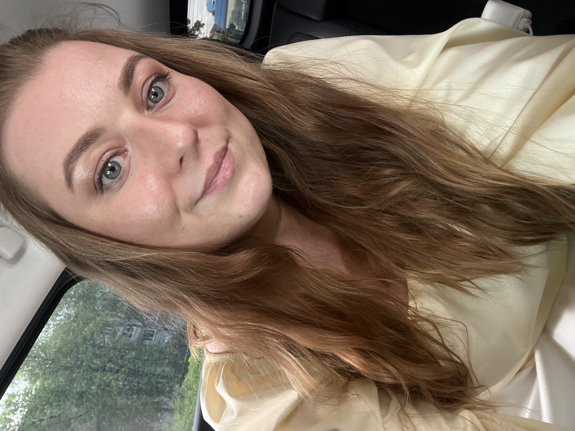 A selfie of a Caucasian woman with long brown hair, light skin, and blue eyes, wearing a light yellow blouse, is taken inside a vehicle. The setting appears to be daytime, judging by the lighting. Visible through the car window are green trees and a building, possibly indicating a residential area. The location is not identifiable as Balabanovo, Russia based on the visible information. The woman is looking directly at the camera with a slight smile. A white object and dark panel are visible in the background within the car.