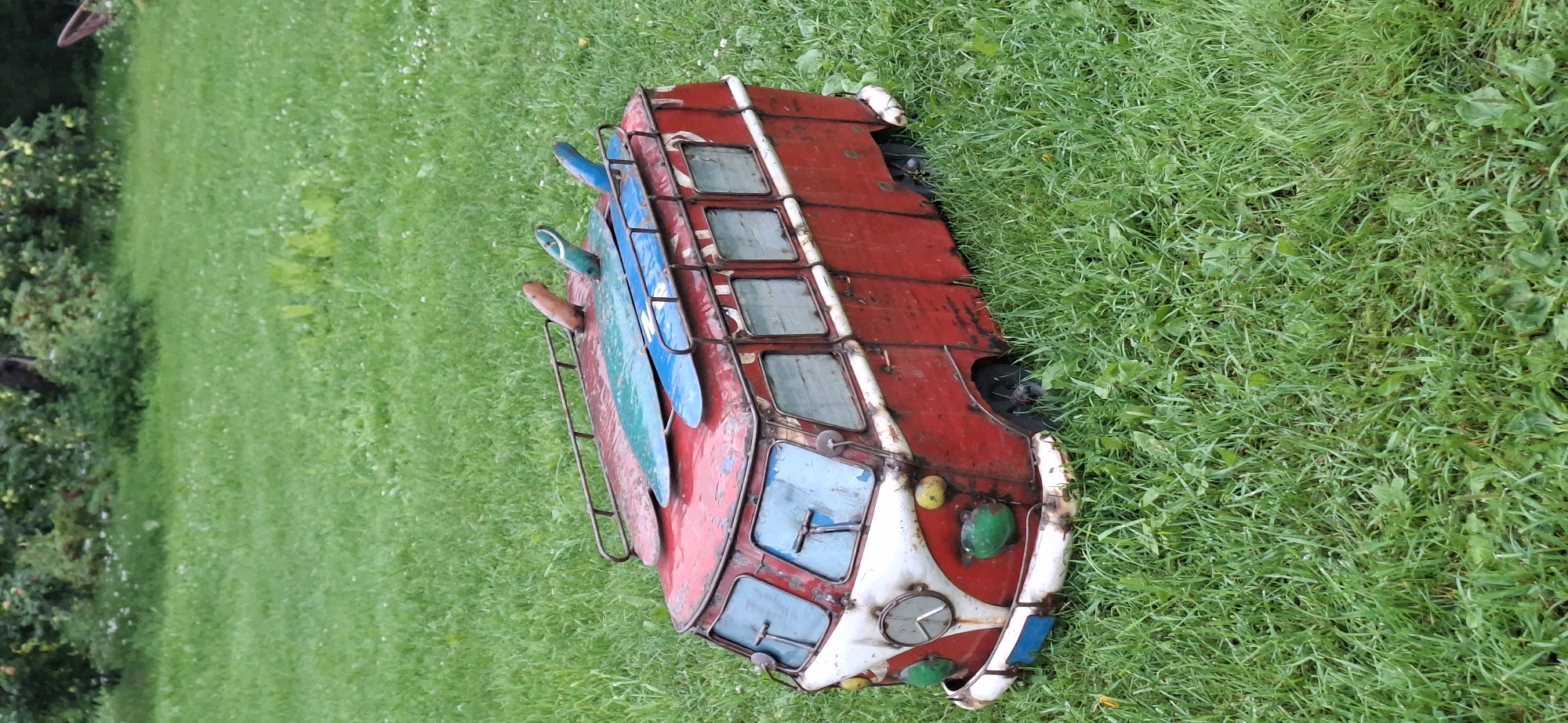 A vintage-style, red and white metal camper van replica, adorned with three surfboards on its roof rack, sits in a grassy outdoor area. The replica van has a classic Volkswagen Type 2 silhouette, complete with round headlights and a prominent front grille. The surfboards are colored blue and green, with one partially obscured by another. The van is resting on a patch of green grass, with some scattered dandelions visible. The background consists of more green grass and blurred foliage, suggesting a natural outdoor setting. The image is taken from a slightly elevated angle, looking down at the van.