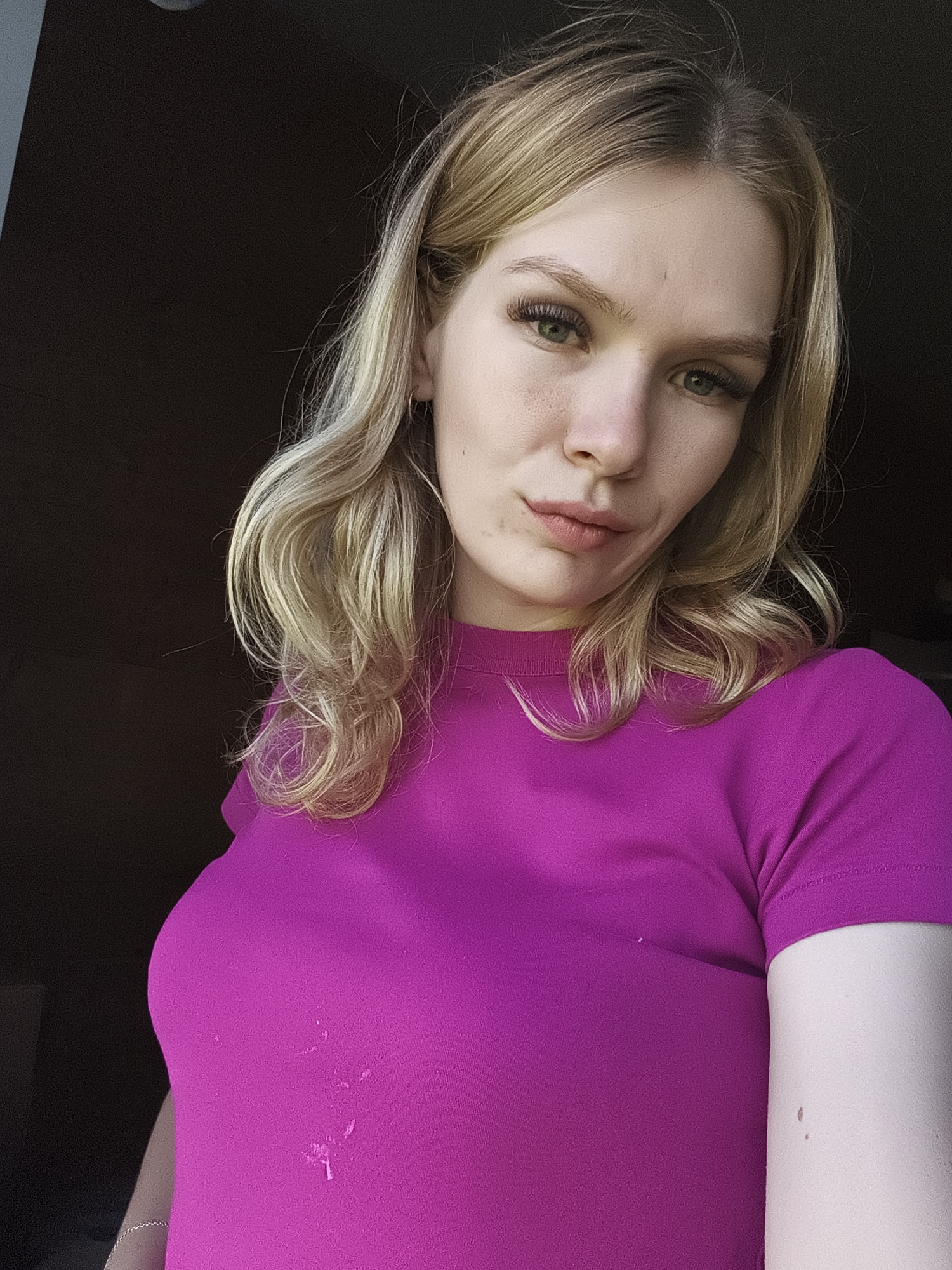 A young woman with blonde hair and green eyes is shown from the chest up, wearing a bright pink shirt. She has long eyelashes and her hair is styled in loose waves. The background is dark and out of focus, suggesting an indoor setting with limited light. There are some light-colored stains on the front of her shirt. The location is not discernible from the image itself; no urban environment or specific geographic features are visible, and therefore no connection to Русский Вожой, Russia can be made.