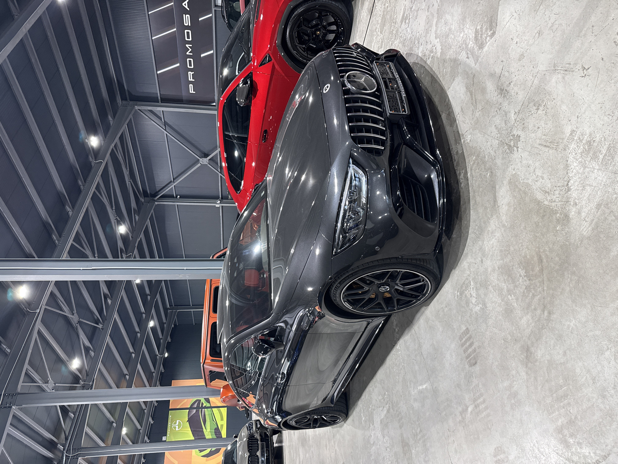 The image depicts an indoor car showroom in Plovdiv, Bulgaria. The foreground features a dark gray Mercedes-AMG GT 4-Door Coupe, angled towards the right. Its interior is visible, revealing red leather seats. Behind it, a bright red Lamborghini Urus is parked, also angled to the right. Further in the background, an orange Mercedes-Benz G-Class is partially visible, and a silver BMW X6 M is seen to the left of the Mercedes-AMG GT. The showroom has a concrete floor with tire marks and is illuminated by overhead industrial lighting. The background walls are dark gray, with one wall featuring signage that reads "PROMOGALE" in white lettering. The overall scene is a display of luxury vehicles in a commercial setting.