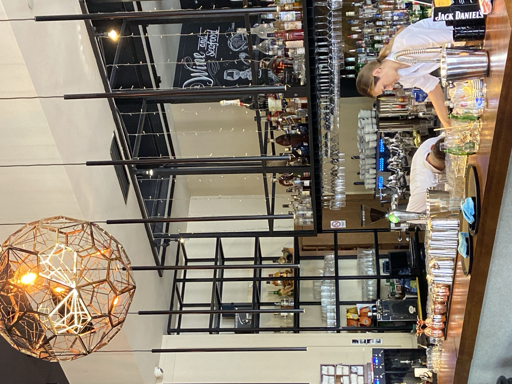 The image captures an interior scene within a bar or restaurant in Budva, Montenegro, featuring a modern industrial aesthetic. The main focus is a long bar counter with two bartenders actively working.
On the right side of the frame, a female bartender, wearing a white shirt and with her hair in a ponytail, is seen mixing a drink behind the wooden bar counter. A large metal shaker and various glasses are around her. Further down the bar, another male bartender, also in a white shirt, is bent over the counter, engaged in preparing drinks or tending to the bar area. Behind them, shelves are stocked with numerous bottles of spirits and liqueurs, along with rows of clean glassware. A "Jack Daniel's" branded item is visible on the counter.
The central and left portions of the image showcase the establishment's decor. A large, ornate, geometric light fixture or chandelier, made of interconnected golden frames, hangs prominently in the foreground on the left, emitting a warm glow. Behind the bar, an open, industrial-style shelving unit extends across the wall, holding more bottles and an array of hanging and stacked glasses. Decorative string lights are draped across these shelves and the ceiling area. A chalkboard sign is visible among the bottles, with stylized white text that reads "Wine and Seafood." Other bar equipment, including coffee machines and beer taps, can be seen behind the counter. A "no smoking" sign is also visible on one of the shelves. The overall impression is a bustling bar environment with contemporary design.