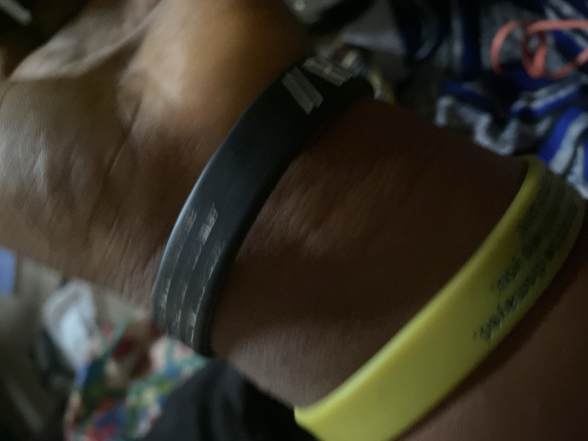 The image shows a close-up of a person's wrist wearing two wristbands. The wristband closer to the hand is black and appears to have some scuff marks and possibly some silver text or design on it, though it is partially obscured. The wristband further down the arm is bright yellow and also appears to have some faint text or markings. The skin tone of the wrist suggests it belongs to a person of color. The background is out of focus but appears to be an indoor environment with some patterned fabric and other objects that are not clearly discernible. The lighting is dim. There is no indication of the location being in Jauro Yinu, Nigeria, as the image only contains a close-up of a wrist with accessories.