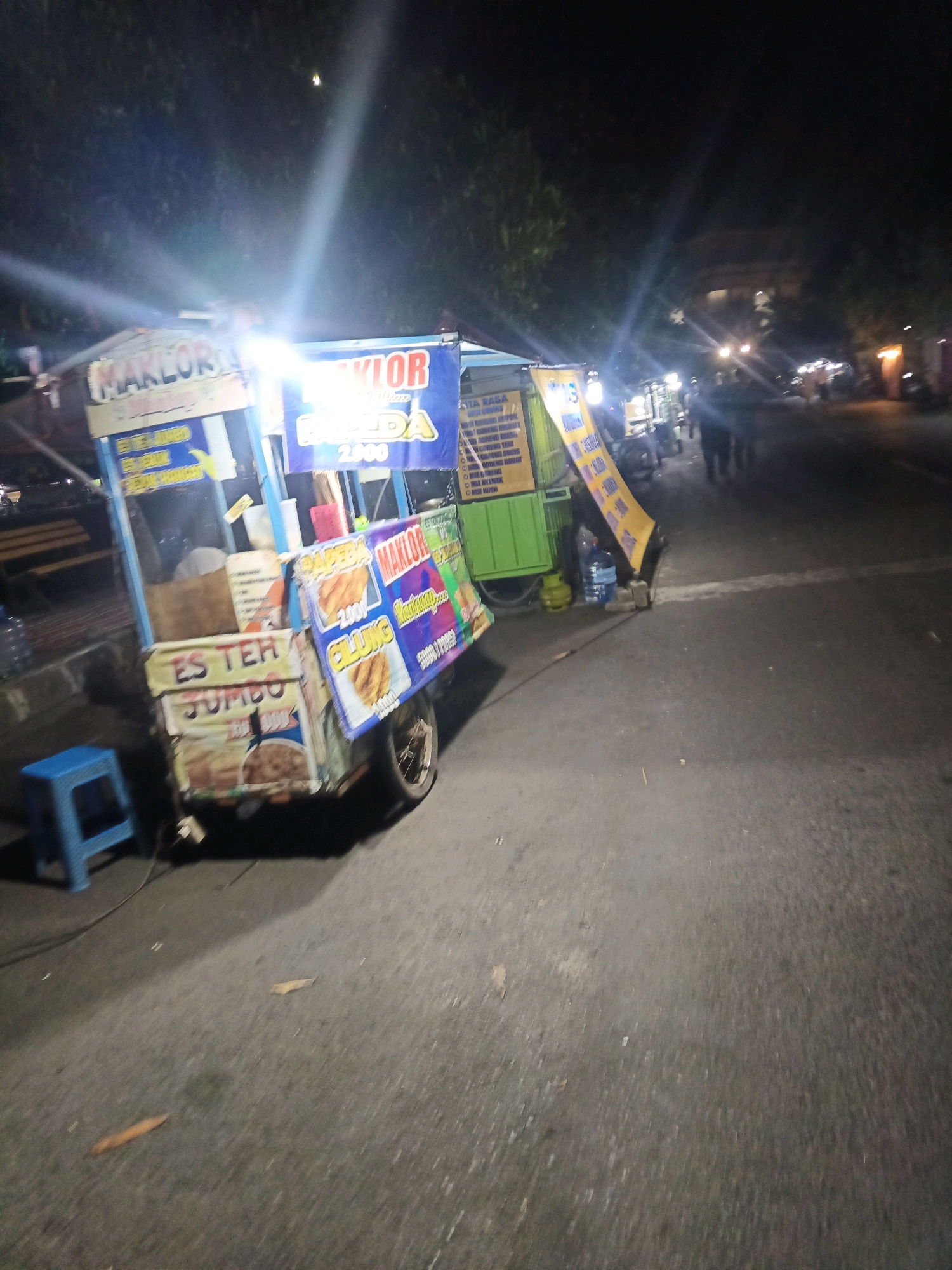 An outdoor, nighttime street scene in Ngasem, Indonesia. The primary subject is a food cart that is illuminated by bright lights. The cart appears to be a mobile vendor, with a blue and white structure and various signs advertising food and drinks. One prominent sign reads "MAKLOR" in large letters. Another sign indicates "ES TER JUMBO". There are also images of fried food items on the cart's signage, suggesting it sells snacks. A blue stool is placed near the cart. In the background, trees and some buildings are visible, with streetlights and vehicle lights suggesting a moderate amount of urban activity. Several blurred figures of people are visible in the mid-ground, walking along the street. The overall impression is of a bustling street food vendor operating at night.