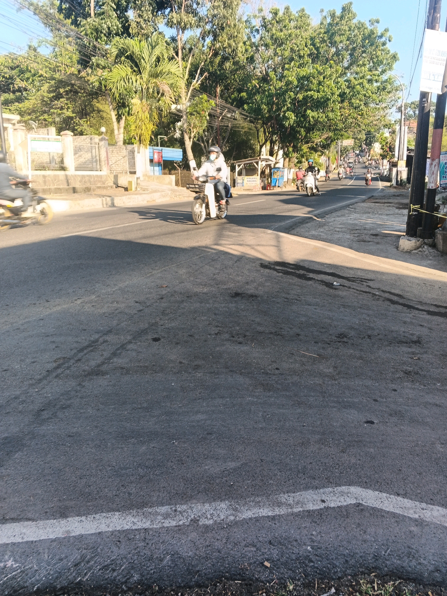The image depicts a street scene in what is identified as Semarang, Indonesia. The road surface is asphalt, with a white line marking the lane. Several motorcycles are visible. In the foreground, a motorcycle with two riders, one driving and one as a passenger, moves away from the camera. Both riders are wearing helmets and masks. The driver wears a white jacket, and the passenger wears dark clothing. To the left, another motorcycle is blurred due to motion, with a rider in a light-colored shirt. Further down the road, a procession of motorcycles is heading uphill. On the right side of the road, there are utility poles with yellow tape tied around them. Behind the poles, a white sign with text is partially visible. The left side of the road features a low stone wall and a building with a fenced entrance, surrounded by trees. The sky is clear and blue, and the lighting suggests it is daytime, likely late afternoon given the long shadows cast by the trees and poles.