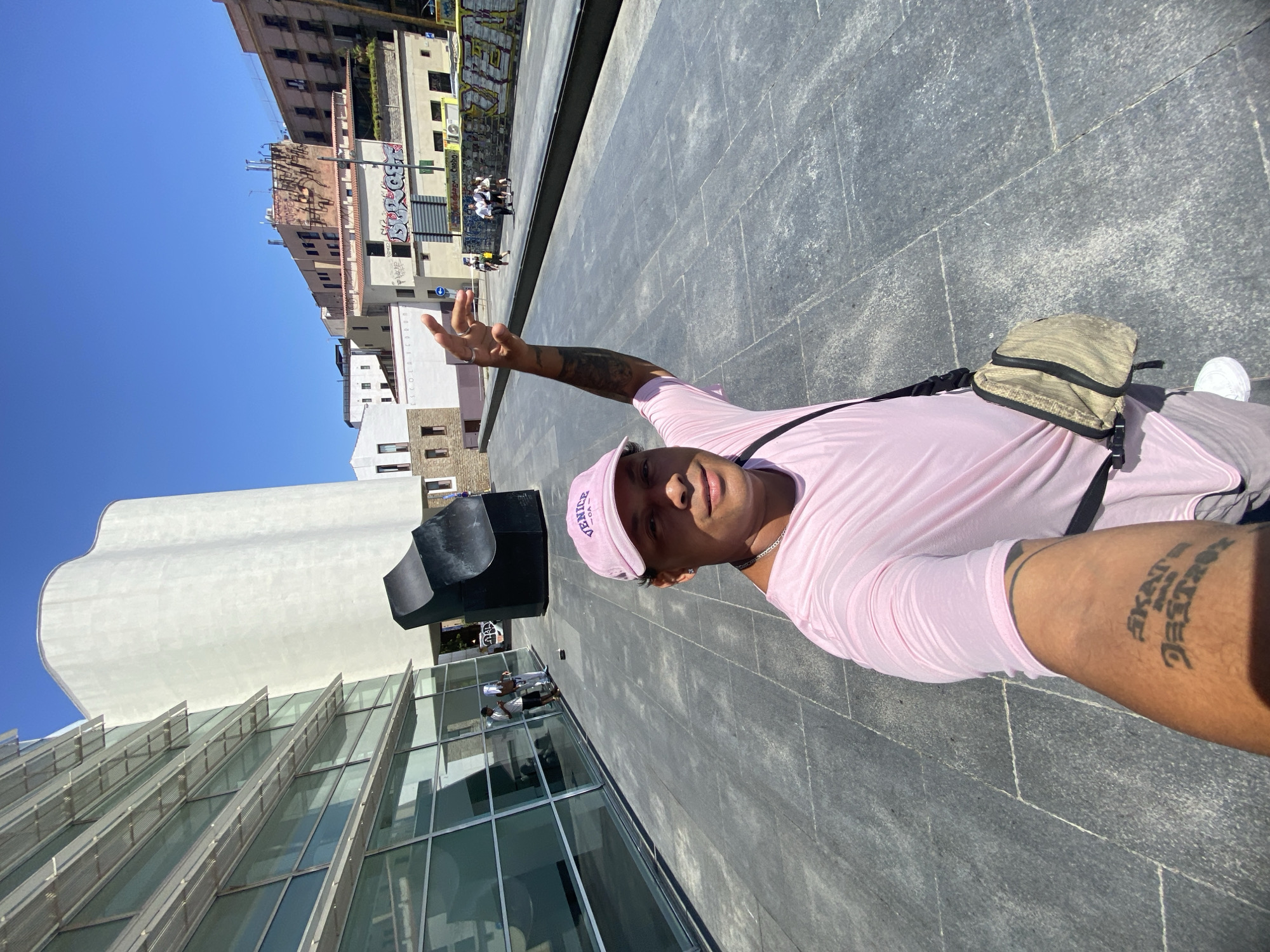 A person is taking a selfie outdoors on a sunny day. They are wearing a pink t-shirt, a pink baseball cap with "VENICE" embroidered on it, and white sneakers. A crossbody bag with a beige and black pattern is slung over their shoulder. The person has tattoos on their arm. They are standing on a paved area with a large, modern building with glass walls and a distinctive wavy white roof in the background. To the left of the modern building, older buildings with graffiti are visible. In the distance, a few people are seated on benches. The sky is clear and blue, suggesting it is daytime. The scene appears to be a public square or plaza.
