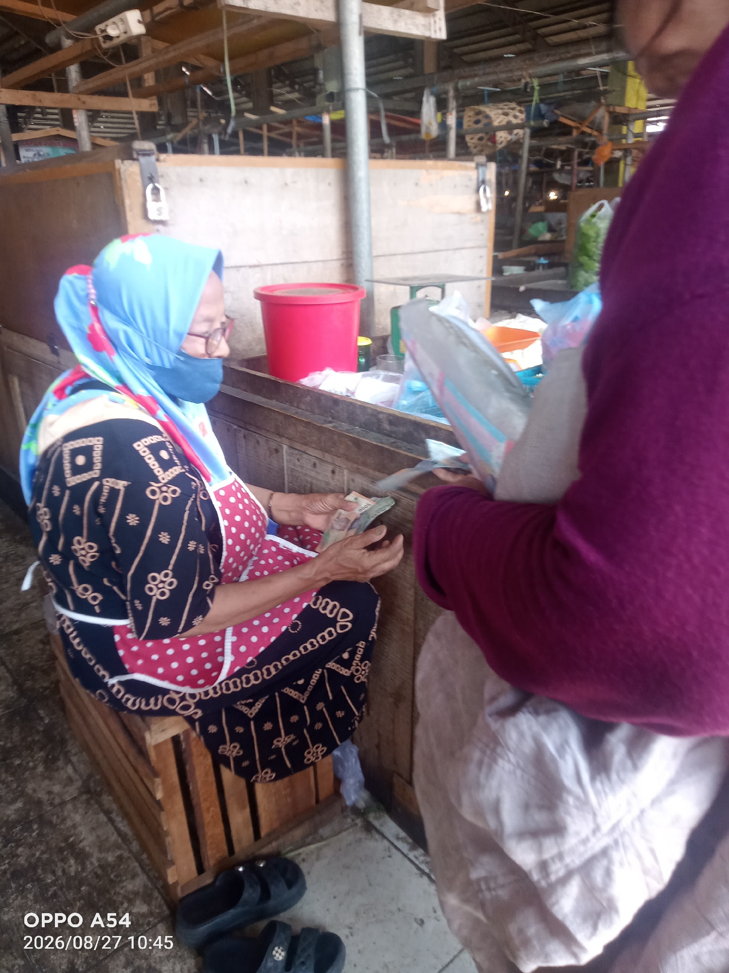 A woman wearing a blue hijab and red polka dot apron sits on a wooden crate at a market stall in Jambi, Indonesia. She is counting money in her hands. Another person, wearing a purple long-sleeved shirt, stands to her right, holding a large object wrapped in plastic. The stall appears to be made of wood and has shelves in the background. The floor is concrete. The image is dated 2026/08/27.