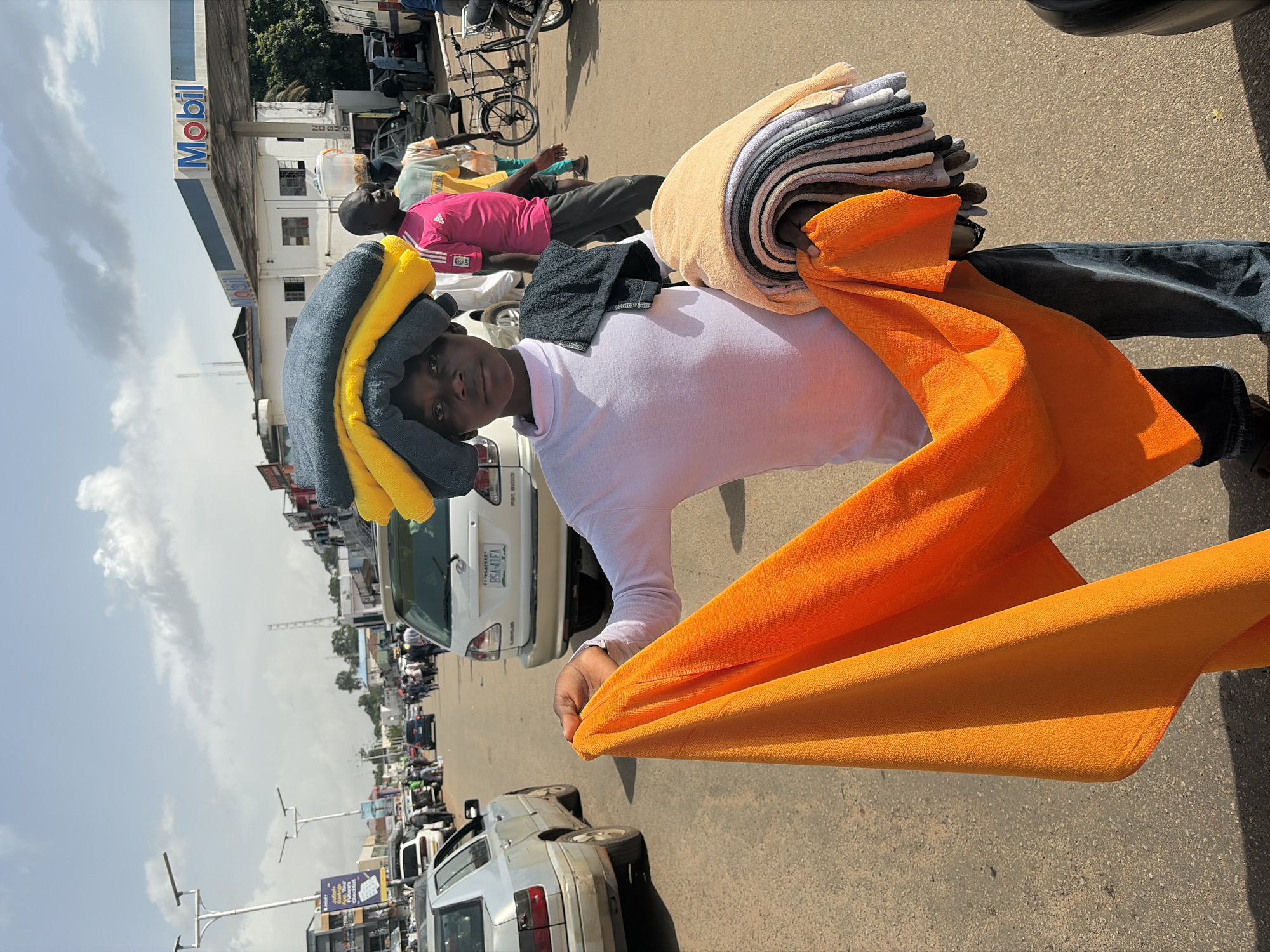 A young person carries several folded towels on their head and arm in a street market environment in Jos, Nigeria. The individual, who appears to be of Nigerian descent, is wearing a white long-sleeved shirt and blue jeans. They are holding a large orange towel in their left hand, while a stack of smaller, multi-colored towels is held in their right arm. The background shows a street with several vehicles, including cars and a motorcycle. A Mobil gas station is visible in the distance, along with various buildings and streetlights. The sky is partly cloudy. The scene suggests a bustling urban setting typical of a Nigerian city.