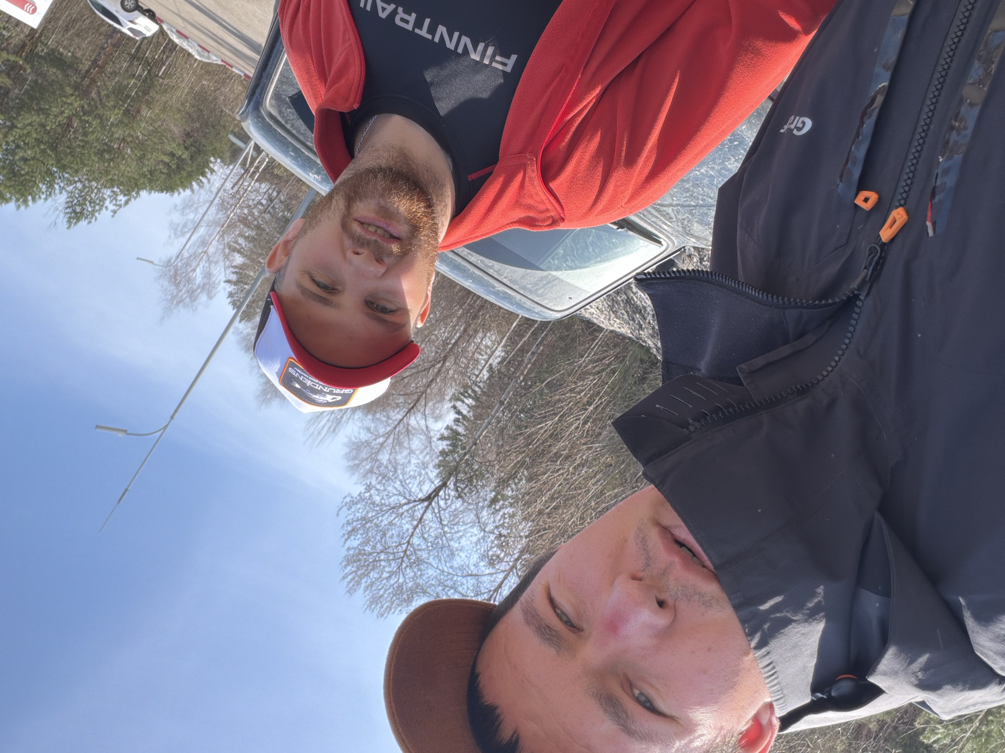 Two men are in the frame. The man on the left is wearing a red jacket over a black shirt with "FINNTRAIL" printed on it, and a red and white baseball cap with a visible logo that reads "GRUNDENS" in white letters and "ENS" in red letters. He has a light beard and is smiling. He is standing behind a car and facing the camera. The man on the right is wearing a black jacket and a brown baseball cap. The camera is pointed upward, indicating that the men are taking a selfie. The environment appears to be outdoors, with trees and a blue sky in the background. A street lamp and a vehicle in the distance are also visible. Without further information, it's not possible to confirm if this photo was taken in Chaikovsky, Russia.