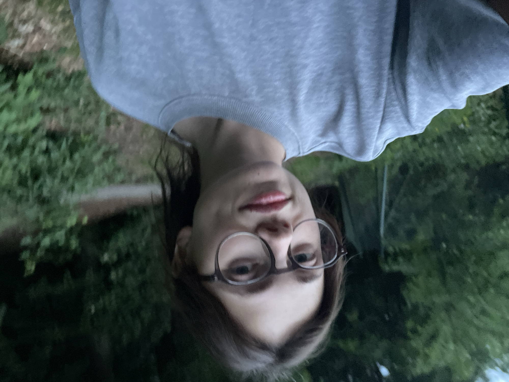 The media file is an upside-down, close-up selfie of a person taken outdoors. The subject is a young individual with medium-length, dark hair, wearing round-framed glasses. They are dressed in a light grey long-sleeved t-shirt or sweatshirt with a ribbed collar visible. The person is looking directly at the camera with a slight, subtle smile.
The background is blurry and consists primarily of green foliage, suggesting a natural outdoor setting such as a park or forest. Some patches of brown or dirt are also visible. The lighting appears somewhat dim, possibly indicating late afternoon, early evening, or an overcast day, contributing to a cool overall tone. No specific landmarks of Karlovy Vary are visible in the image itself, but given the context, the scene is set in a natural area within Karlovy Vary, Czechia. There is no visible text in the image.